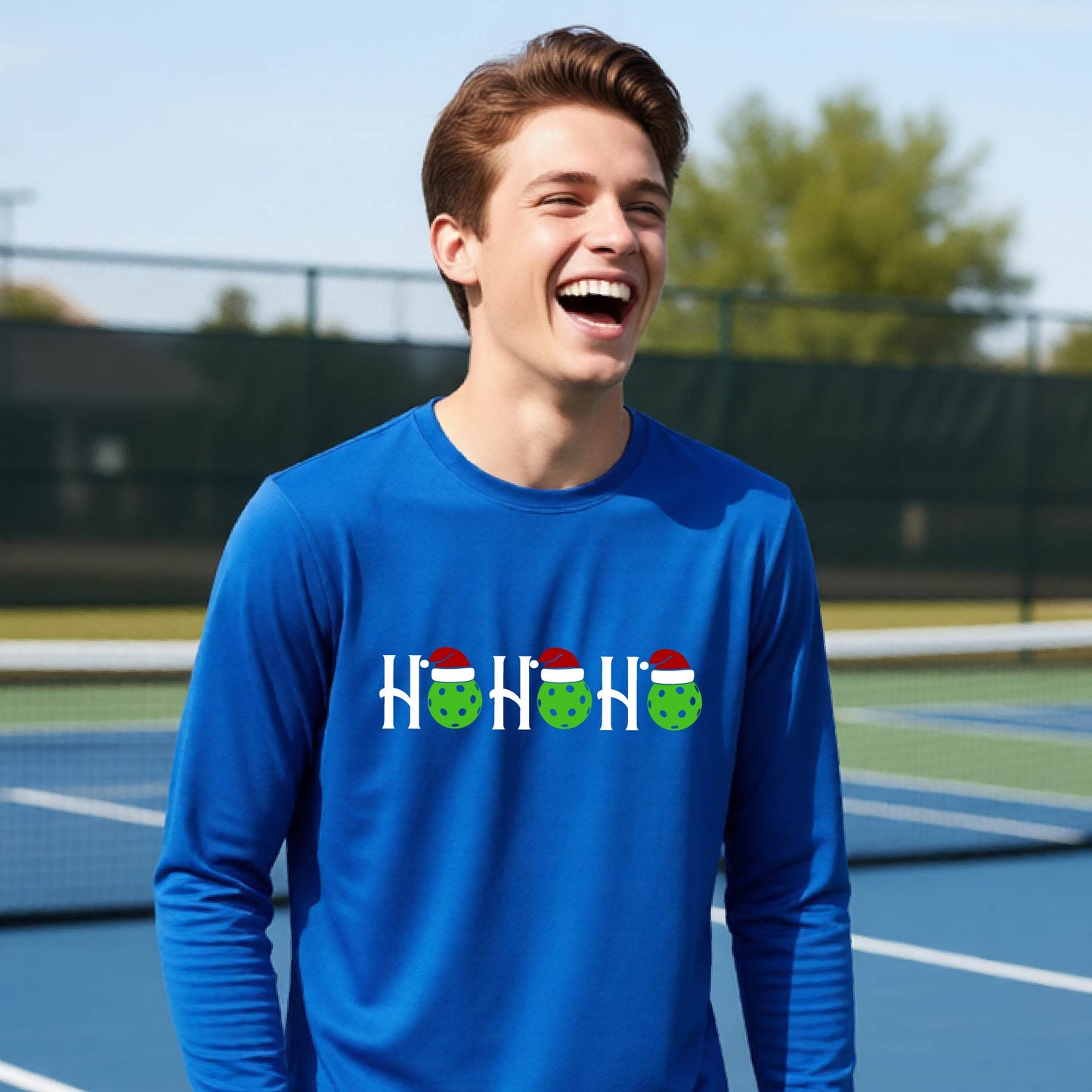 A smiling young person stands on pickleball court wearing a royal blue long-sleeve shirt with a 'HO HO HO' graphic made of green pickleball ornaments wearing red Santa hats.