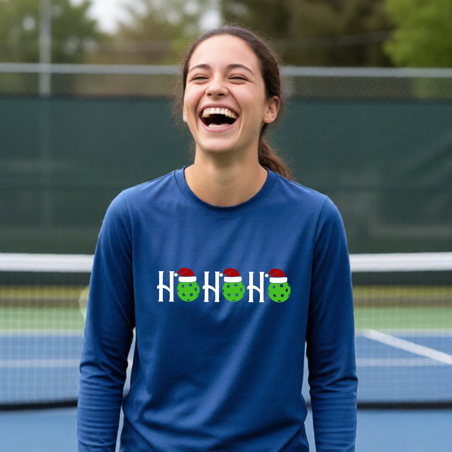 A smiling young person stands on pickleball court wearing a royal blue long-sleeve shirt with a 'HO HO HO' graphic made of green pickleball ornaments wearing red Santa hats.