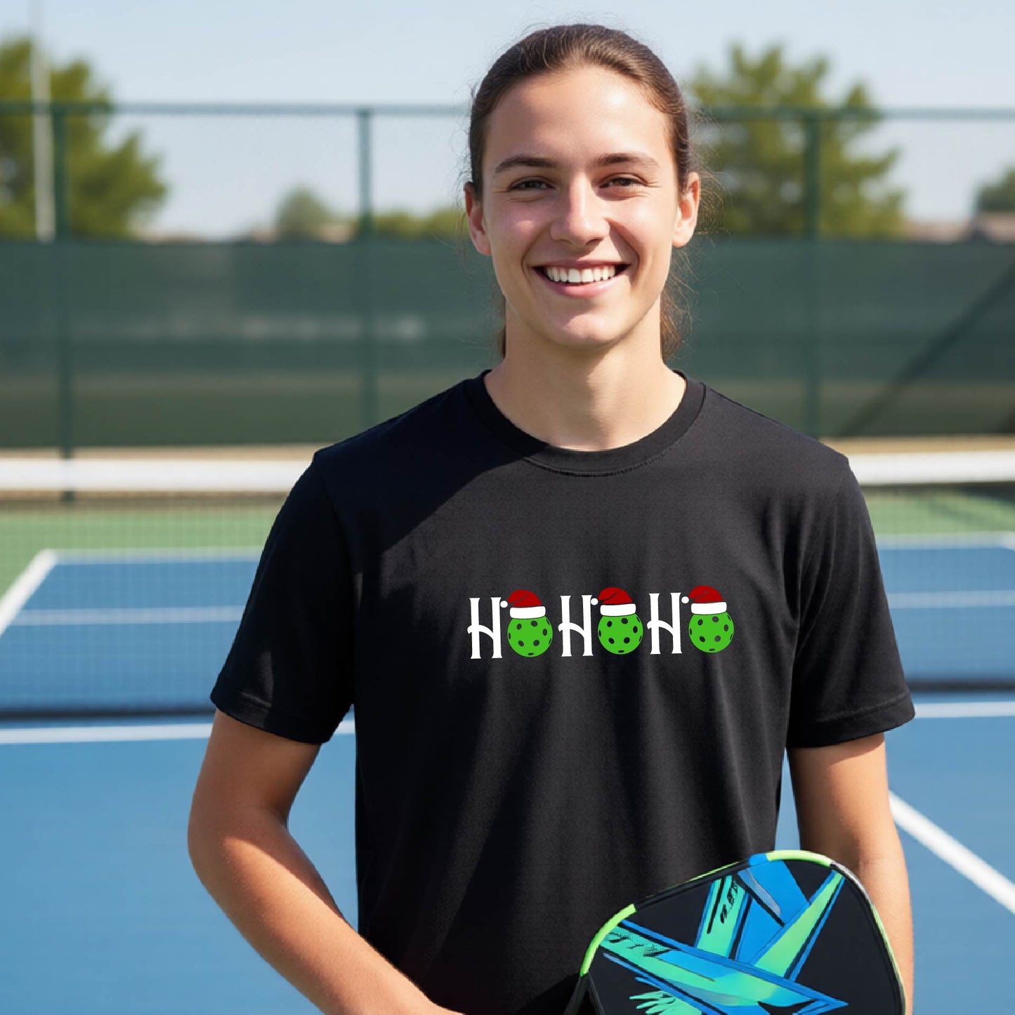 A smiling woman on a blue pickleball court wearing a black t-shirt with a white and green 'HO HO HO' holiday graphic, holding a pickleball paddle.
