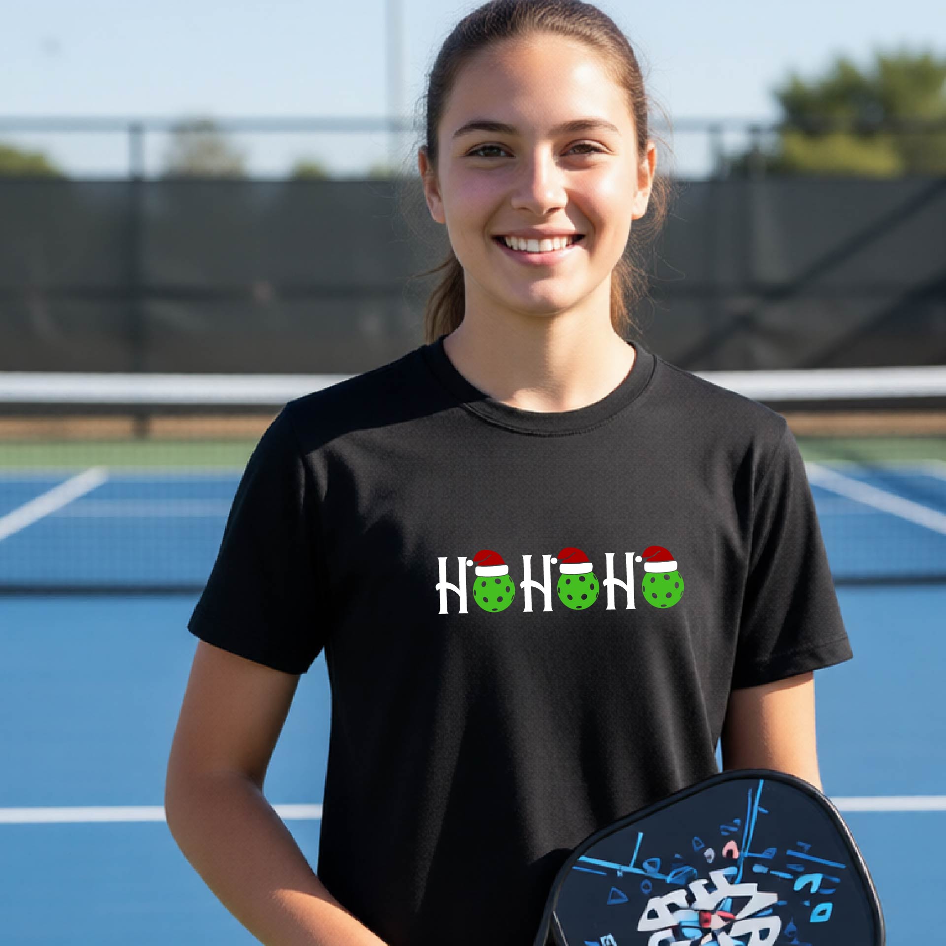 A smiling young woman on a blue pickleball court wearing a black t-shirt with a 'Ho Ho Ho' graphic made of green pickleballs wearing Santa hats. She is holding a black pickleball paddle.