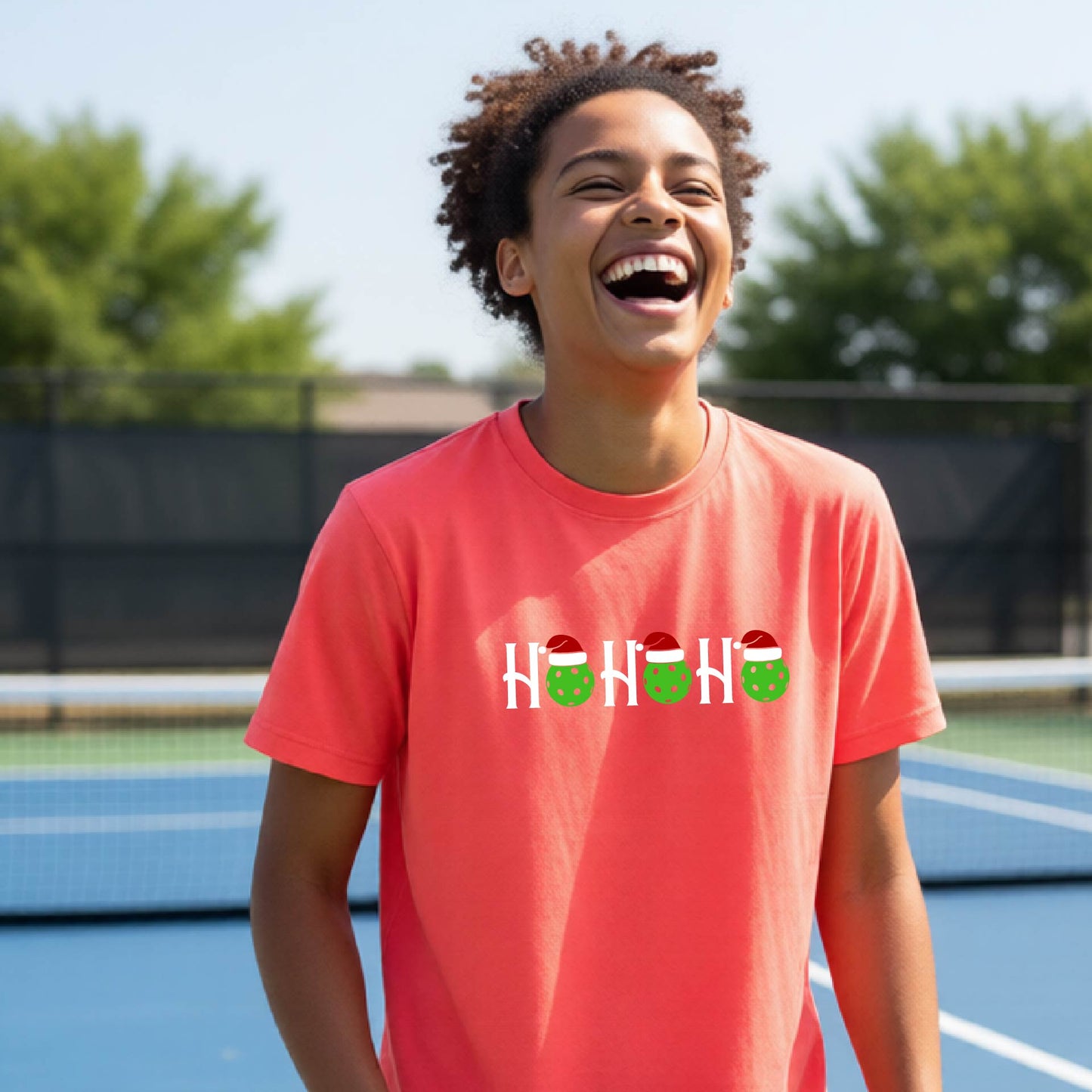 A smiling person standing on a blue pickleball court wearing a coral-pink short-sleeve T-shirt with a white 'HO HO HO' holiday graphic across the chest, with green pickleballs and red Santa hats.