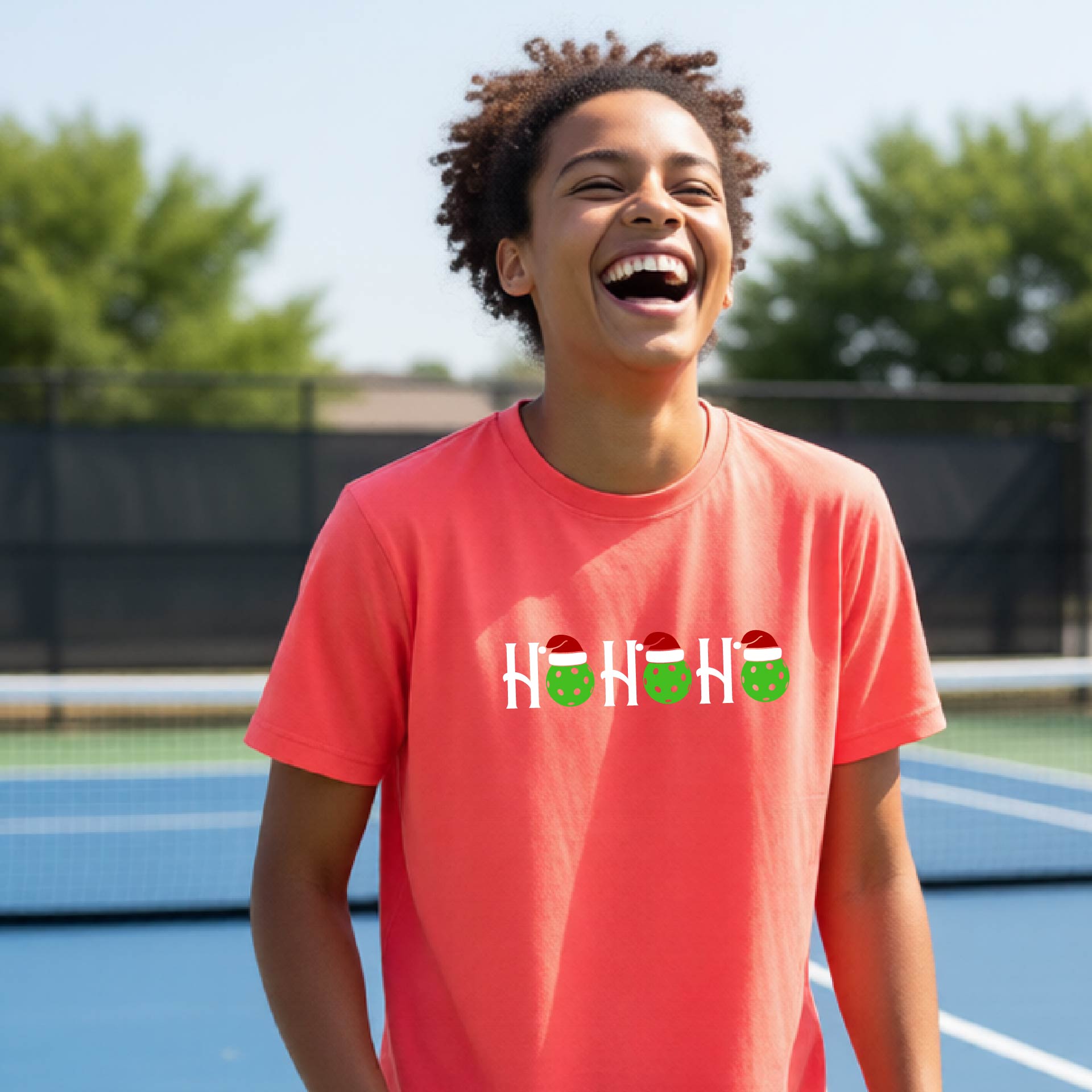 A smiling person standing on a blue pickleball court wearing a coral-pink short-sleeve T-shirt with a white 'HO HO HO' holiday graphic across the chest, with green pickleballs and red Santa hats.