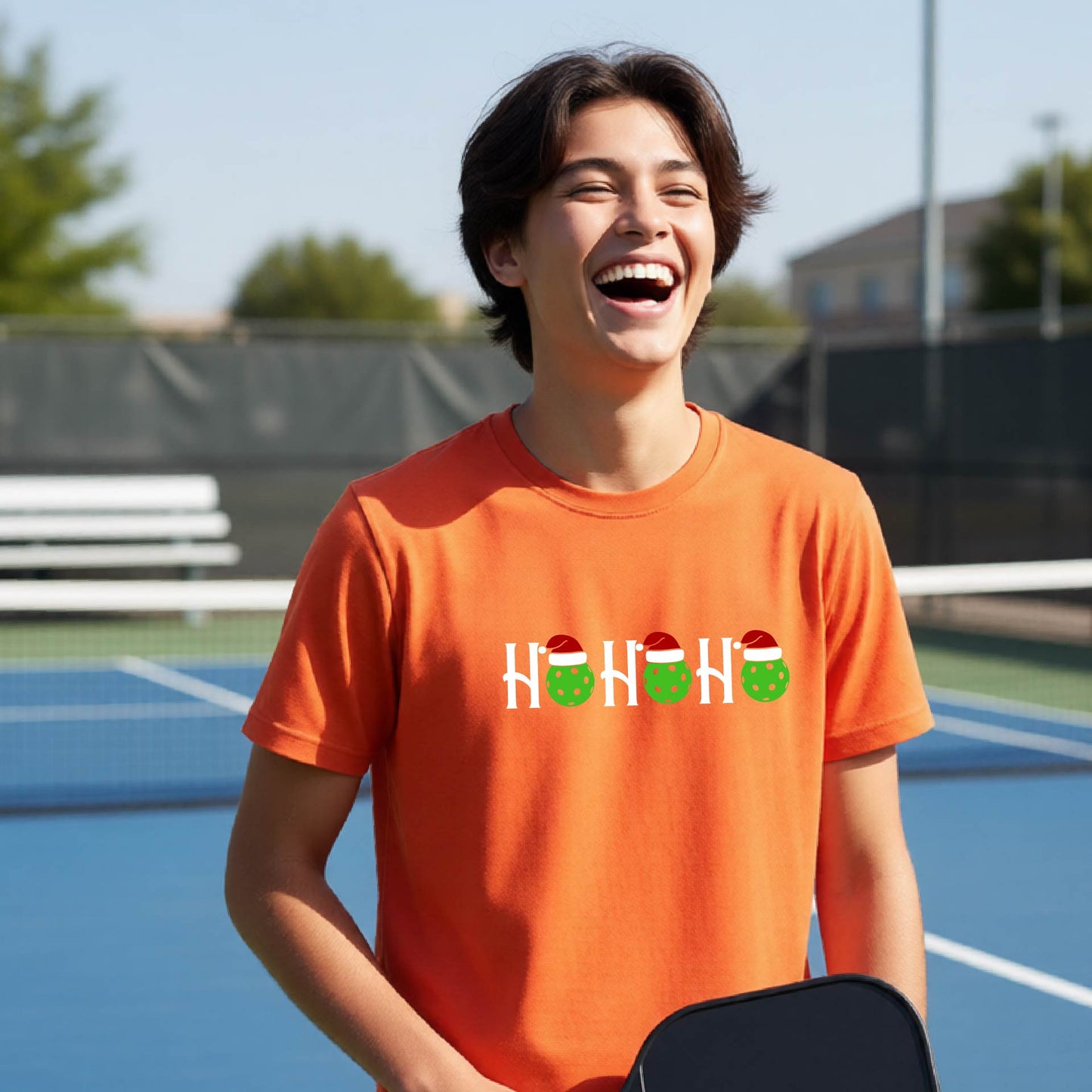 A young man laughing on an outdoor pickleball court wearing a bright orange t-shirt with a white 'HO HO HO' graphic featuring green and white Christmas pickleballs.