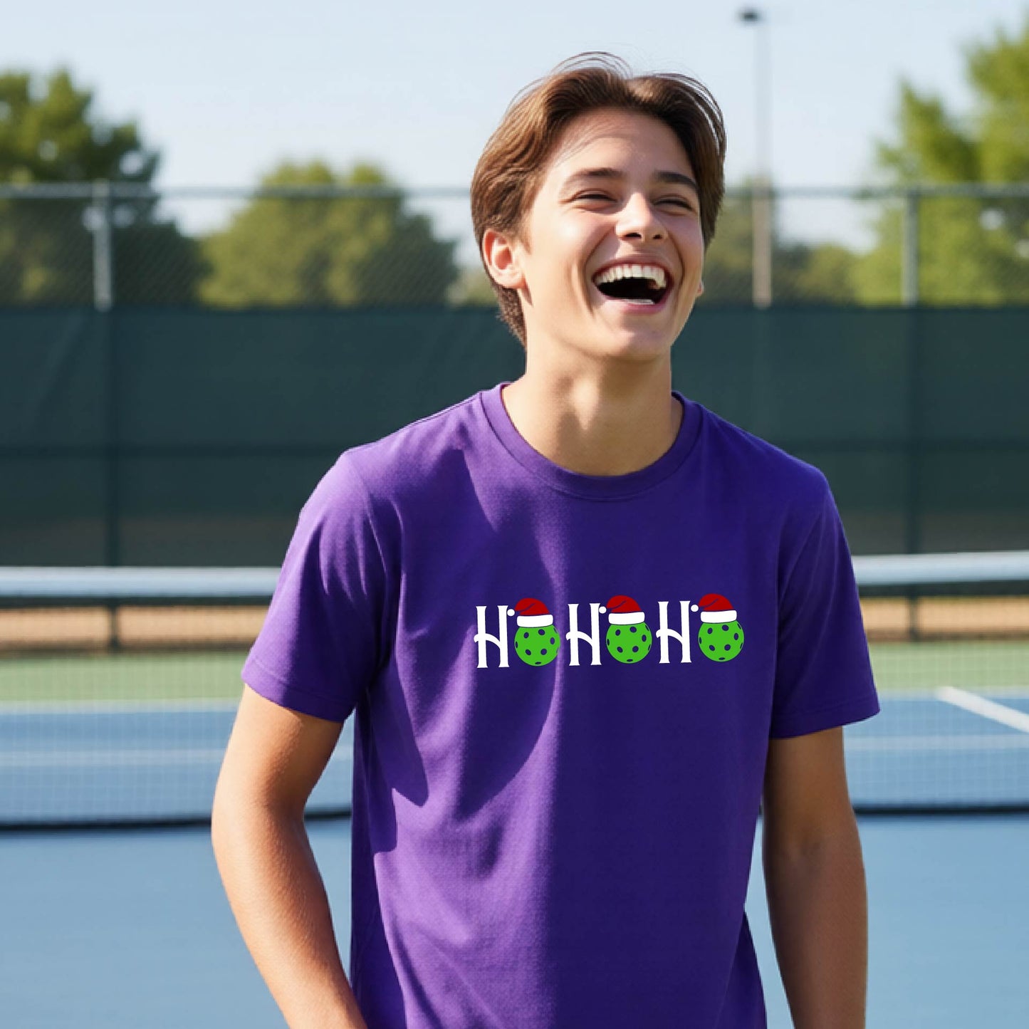 A man laughing on an outdoor pickleball court wearing a purple t-shirt with a 'HO HO HO' graphic made of green pickleballs wearing Santa hats.