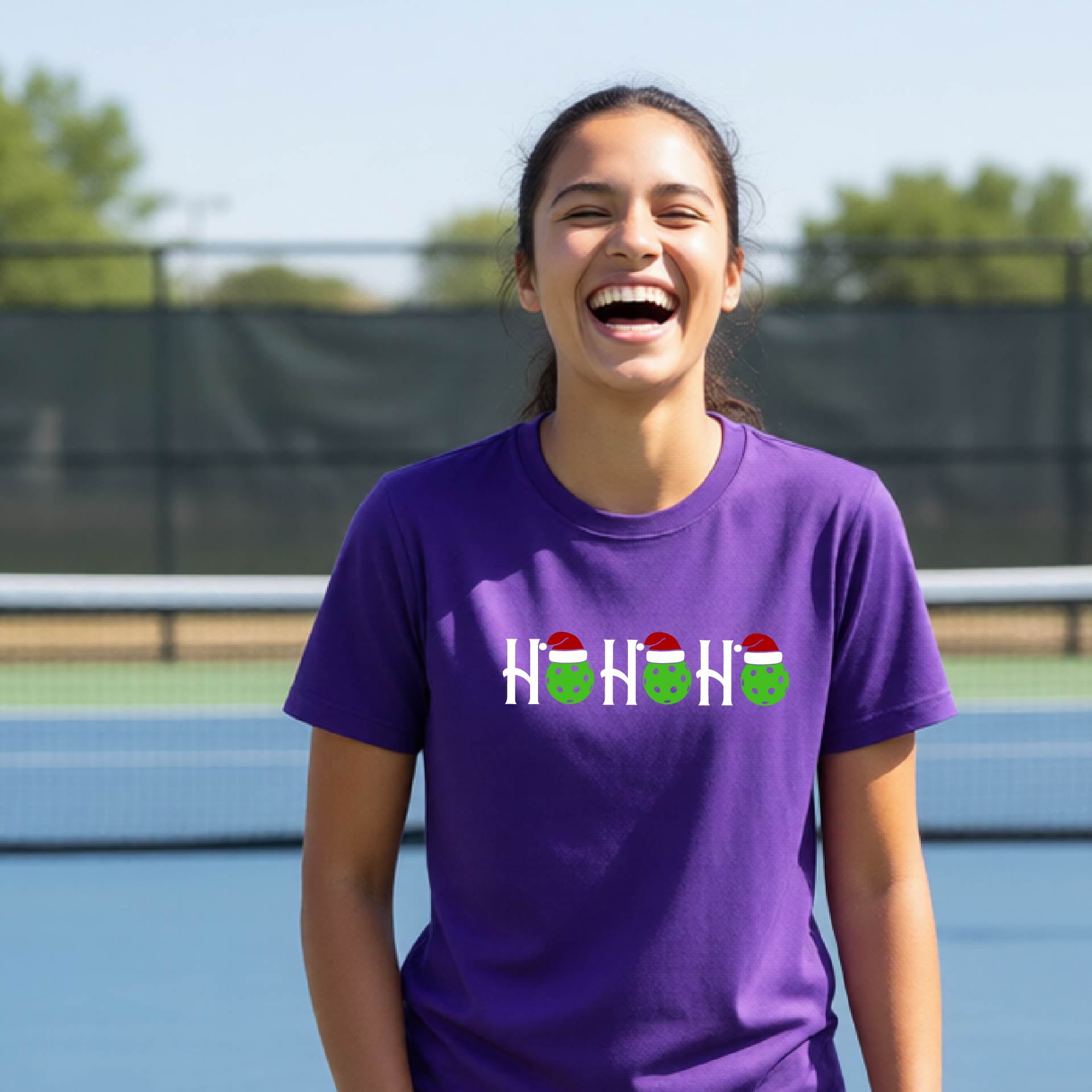 A woman laughing on an outdoor pickleball court wearing a purple t-shirt with a 'HO HO HO' holiday pickleball graphic.