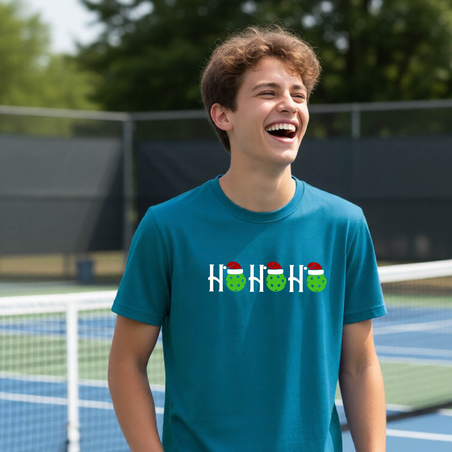 A young man laughing on an outdoor pickleball court wearing a teal t-shirt with a 'HO HO HO' holiday pickleballs graphic.