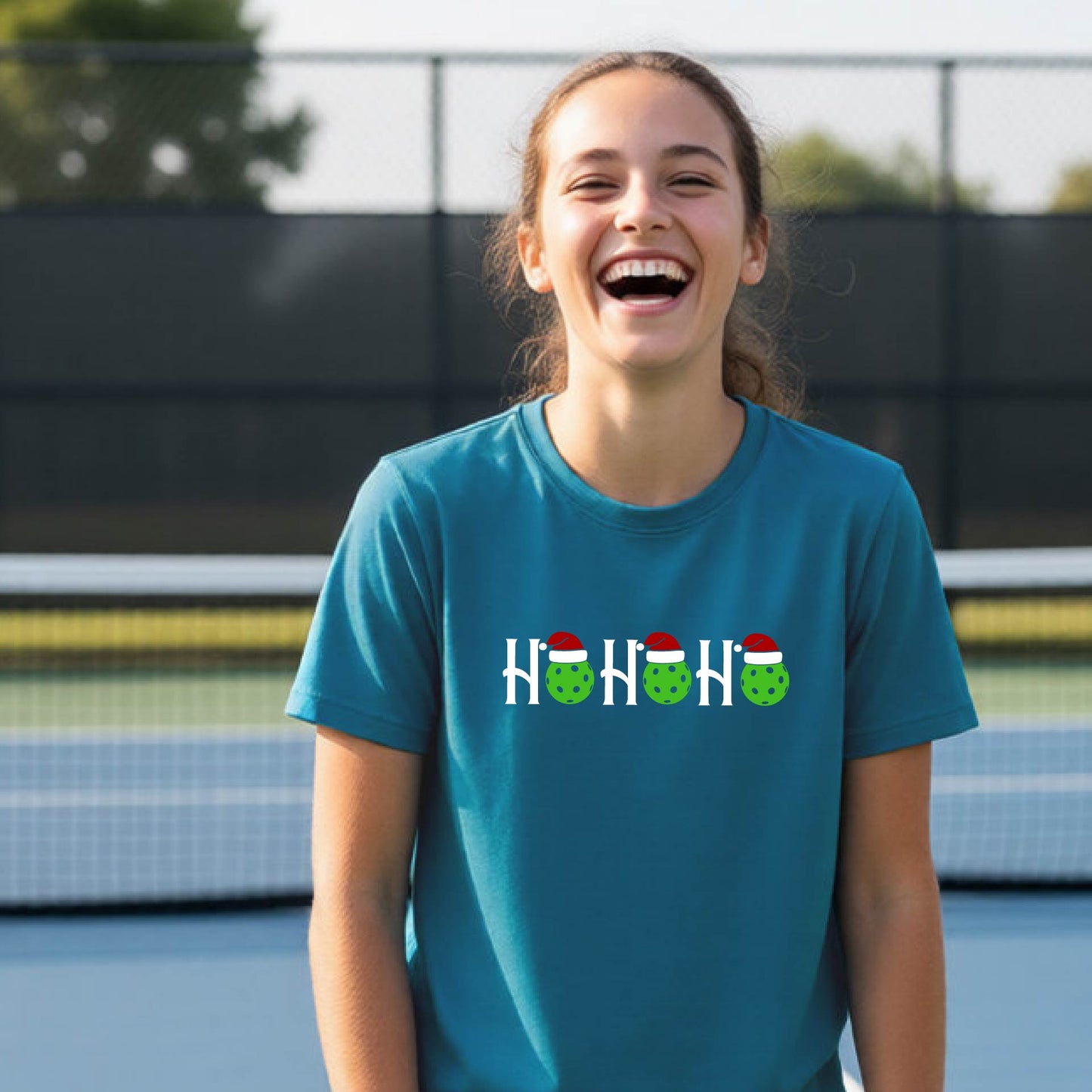 A young woman laughing on an outdoor tennis court wearing a teal blue t-shirt with a graphic that reads 'HO HO HO' in white with green cactus faces wearing red Santa hats.