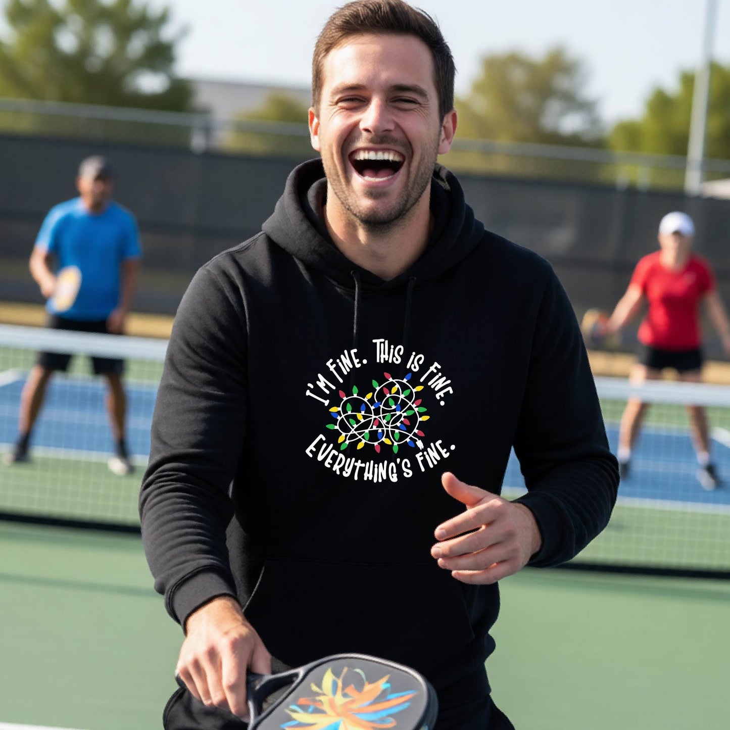 A man smiling while playing pickleball on an outdoor court, wearing a black hoodie with a colorful graphic and the text 'I'M FINE. THIS IS FINE. EVERYTHING'S FINE.
