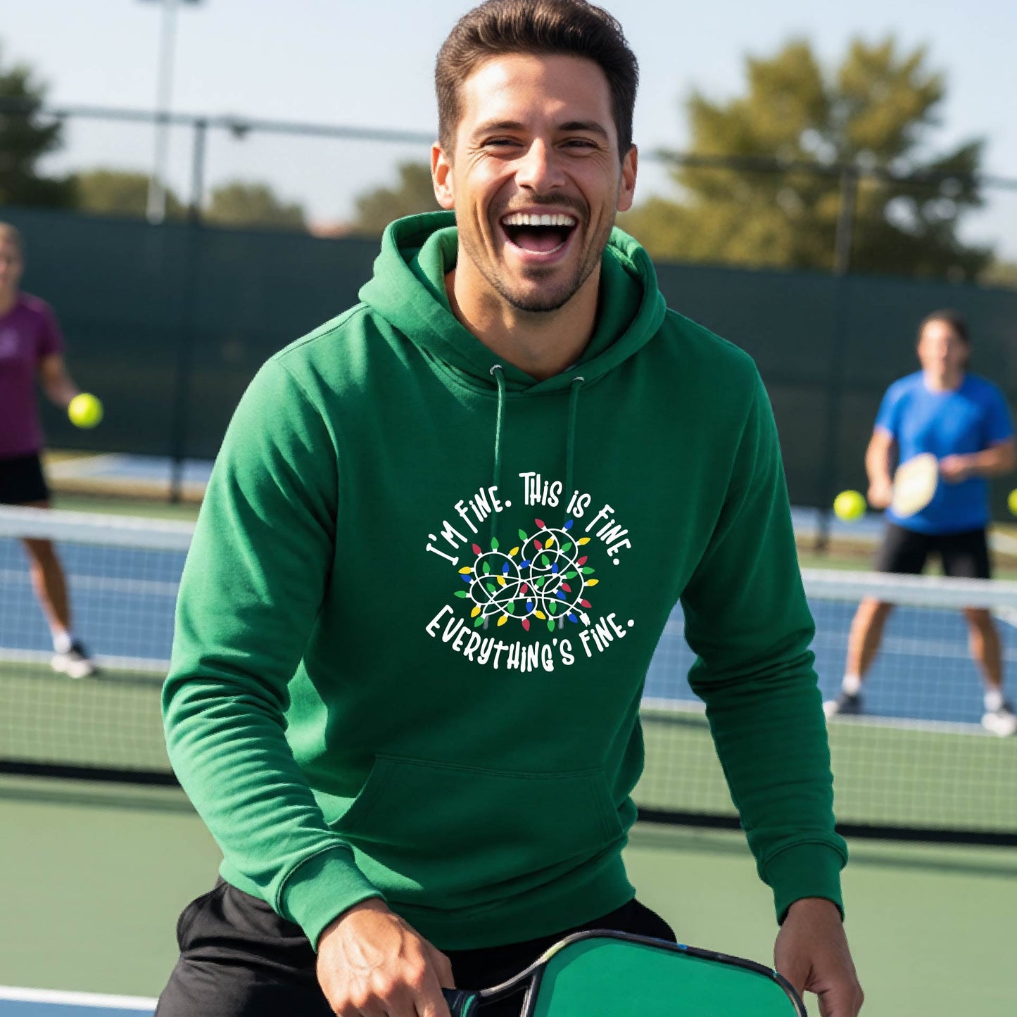 A man smiling while playing pickleball on an outdoor court, wearing a green hoodie with a colorful graphic and the text 'I'M FINE. THIS IS FINE. EVERYTHING'S FINE.