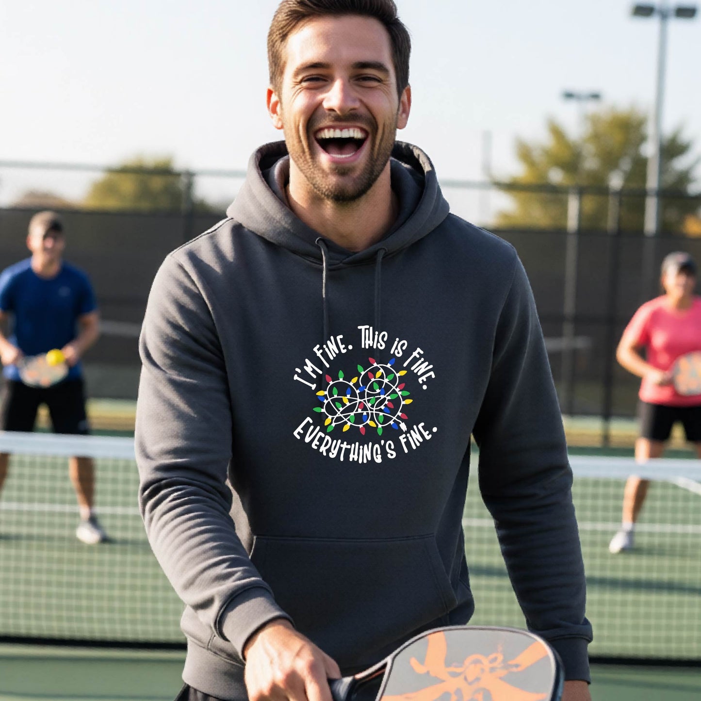 A man smiling while playing pickleball on an outdoor court, wearing a dark gray hoodie with a colorful graphic and the text 'I'M FINE. THIS IS FINE. EVERYTHING'S FINE.