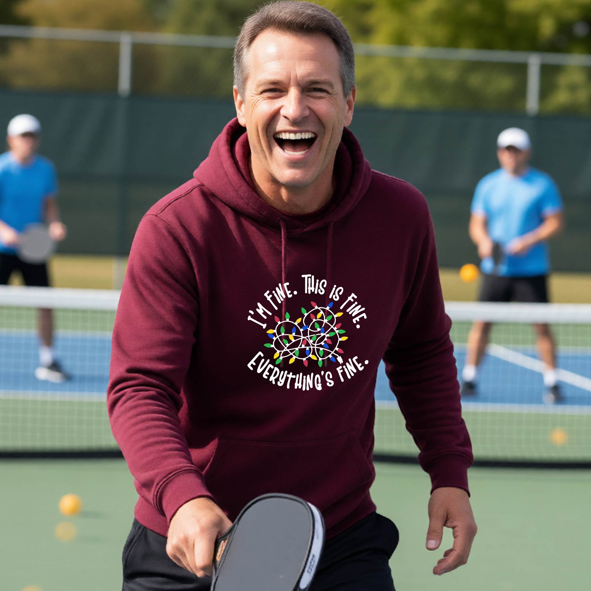 A man smiling while playing pickleball on an outdoor court, wearing a maroon hoodie with a colorful graphic and the text 'I'M FINE. THIS IS FINE. EVERYTHING'S FINE.
