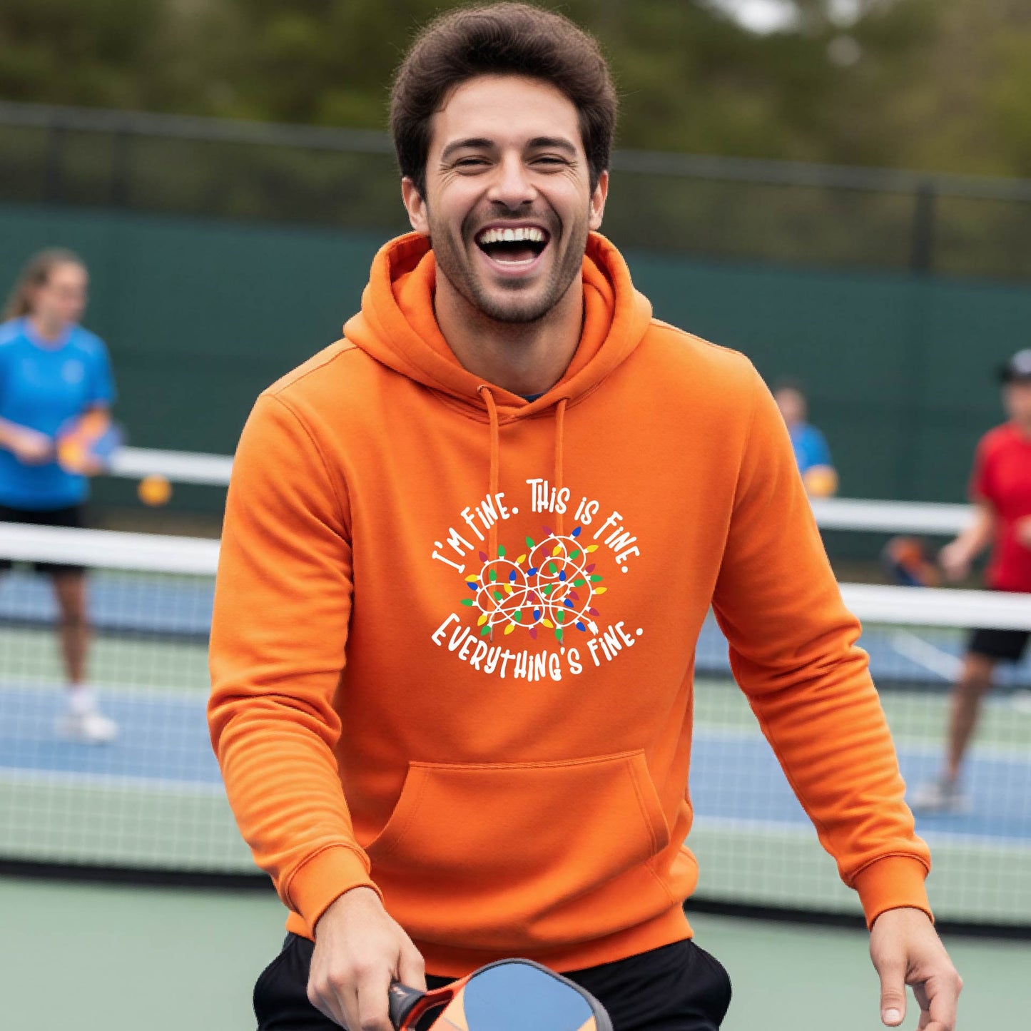 A man smiling while playing pickleball on an outdoor court, wearing an orange hoodie with a colorful graphic and the text 'I'M FINE. THIS IS FINE. EVERYTHING'S FINE.