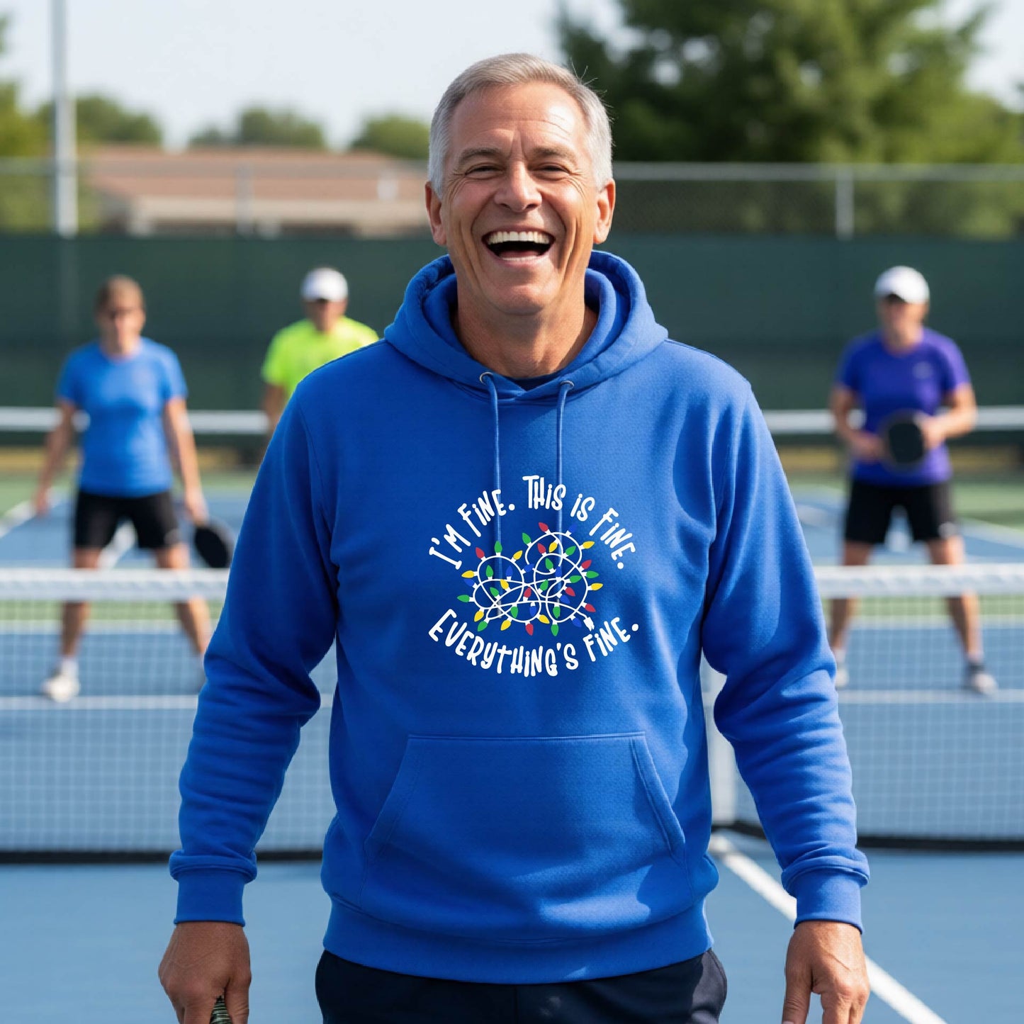 A man smiling while playing pickleball on an outdoor court, wearing a royal blue hoodie  with a colorful graphic and the text 'I'M FINE. THIS IS FINE. EVERYTHING'S FINE.