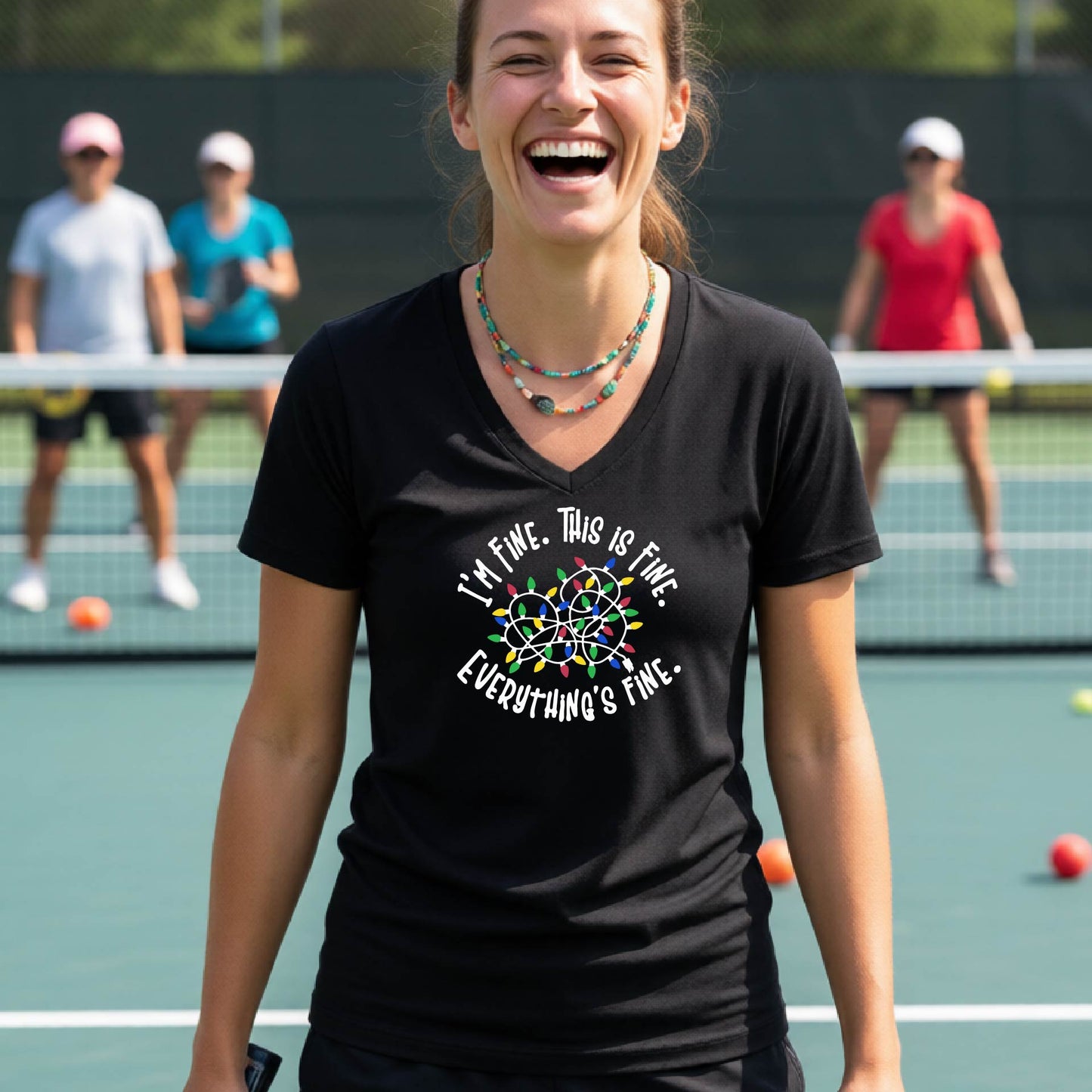A woman smiling on an pickleball court wearing a black short-sleeve t-shirt with a colorful graphic of tangled Christmas lights and the text 'I'M FINE. THIS IS FINE. EVERYTHING'S FINE.'