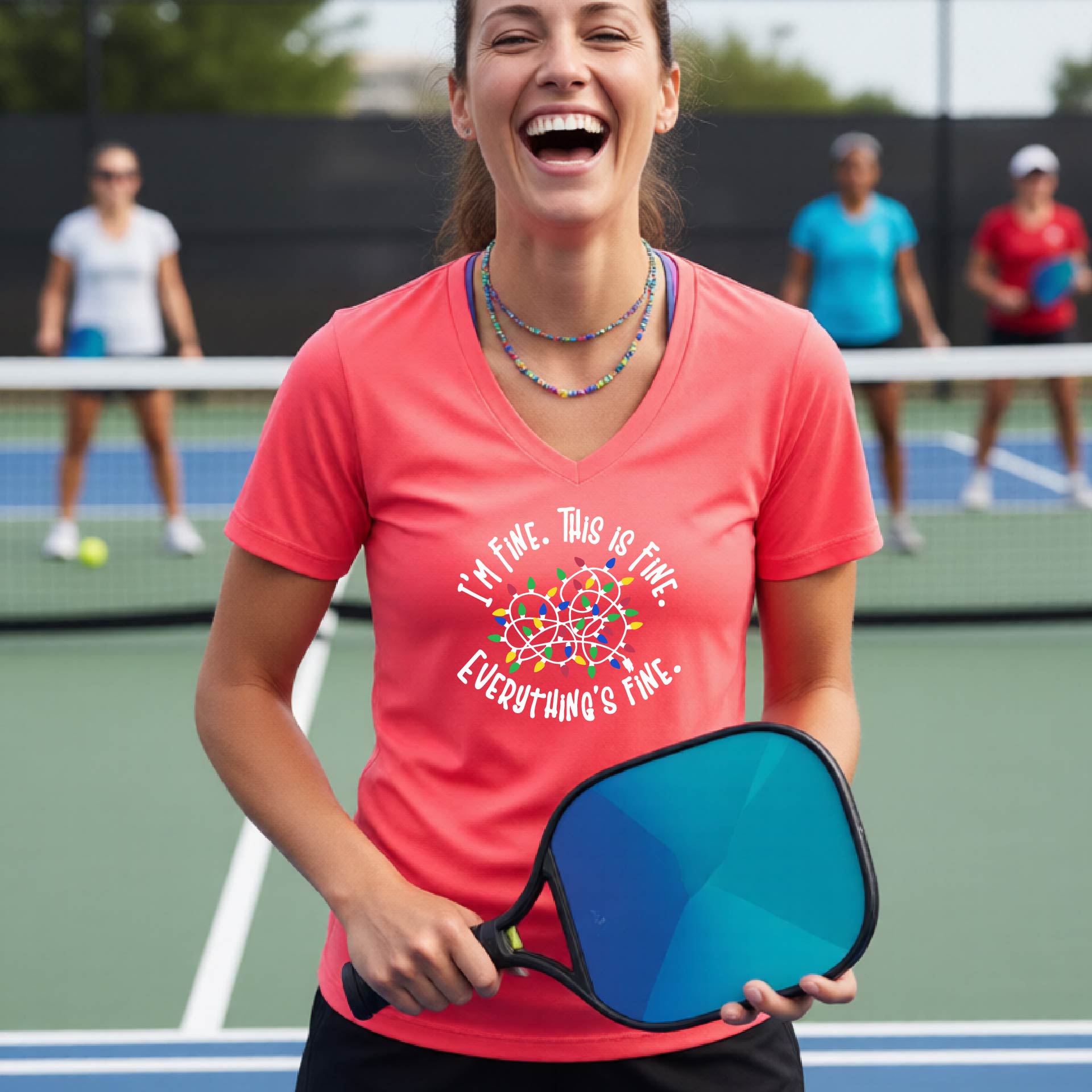 A woman smiling on an pickleball court wearing a coral pink short-sleeve t-shirt with a colorful graphic of tangled Christmas lights and the text 'I'M FINE. THIS IS FINE. EVERYTHING'S FINE.'