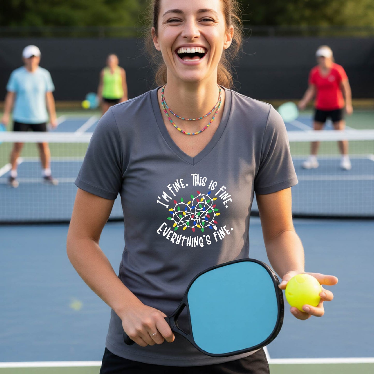 A woman smiling on an pickleball court wearing a dark gray short-sleeve t-shirt with a colorful graphic of tangled Christmas lights and the text 'I'M FINE. THIS IS FINE. EVERYTHING'S FIne.'