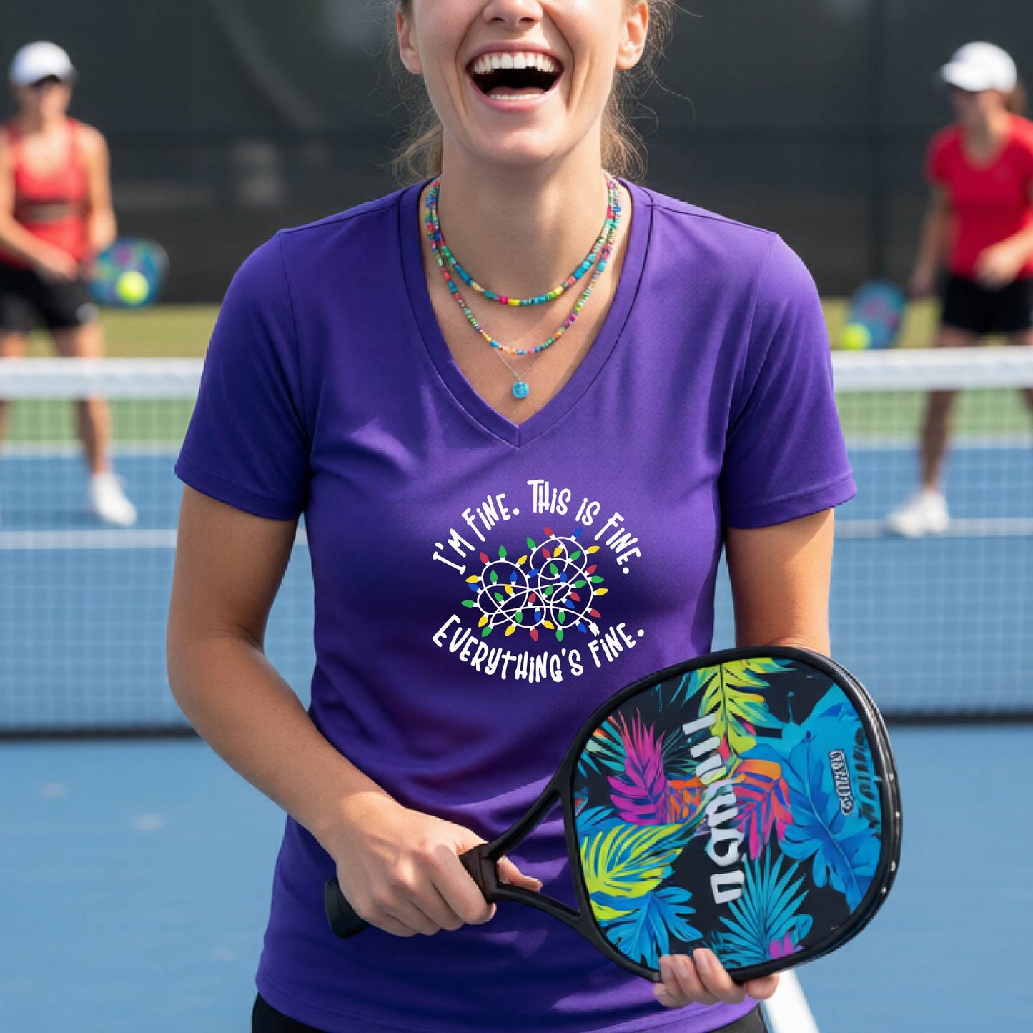A woman smiling on an pickleball court wearing a purple short-sleeve t-shirt with a colorful graphic of tangled Christmas lights and the text 'I'M FINE. THIS IS FINE. EVERYTHING'S FINE.'