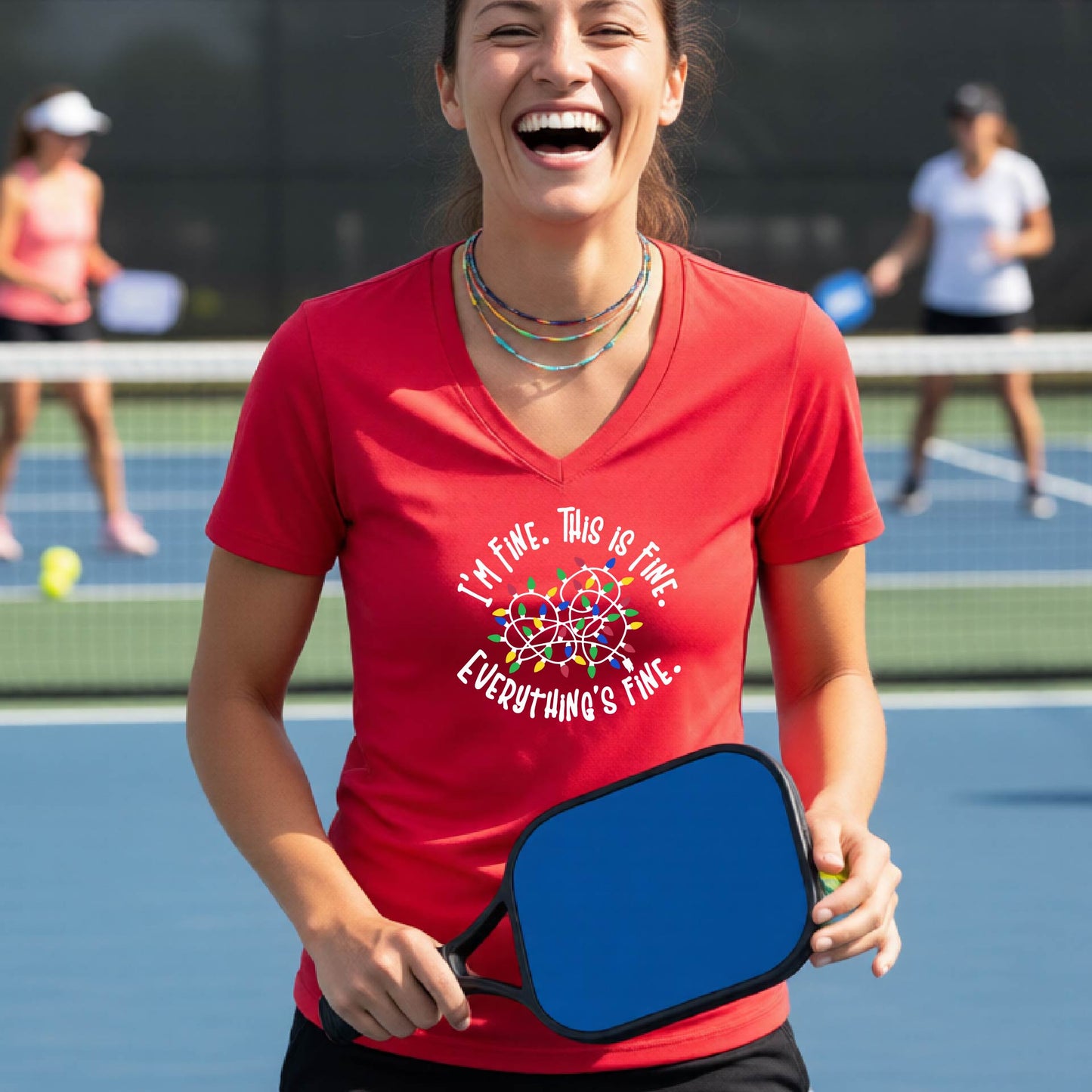 A woman smiling on an pickleball court wearing a red short-sleeve t-shirt with a colorful graphic of tangled Christmas lights and the text 'I'M FINE. THIS IS FINE. EVERYTHING'S FINE.'