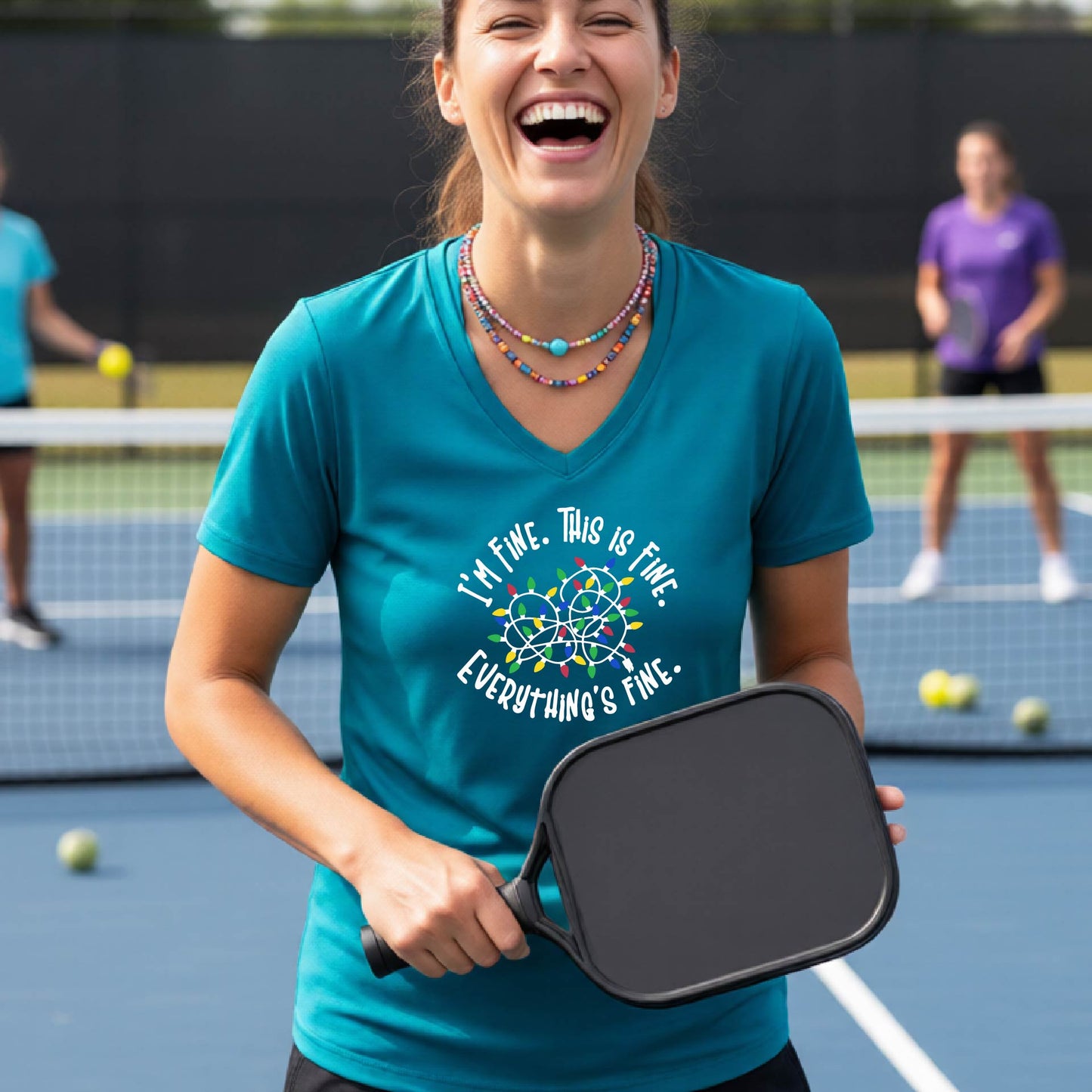 A woman smiling on an pickleball court wearing a teal short-sleeve t-shirt with a colorful graphic of tangled Christmas lights and the text 'I'M FINE. THIS IS FINE. EVERYTHING'S FINE.'
