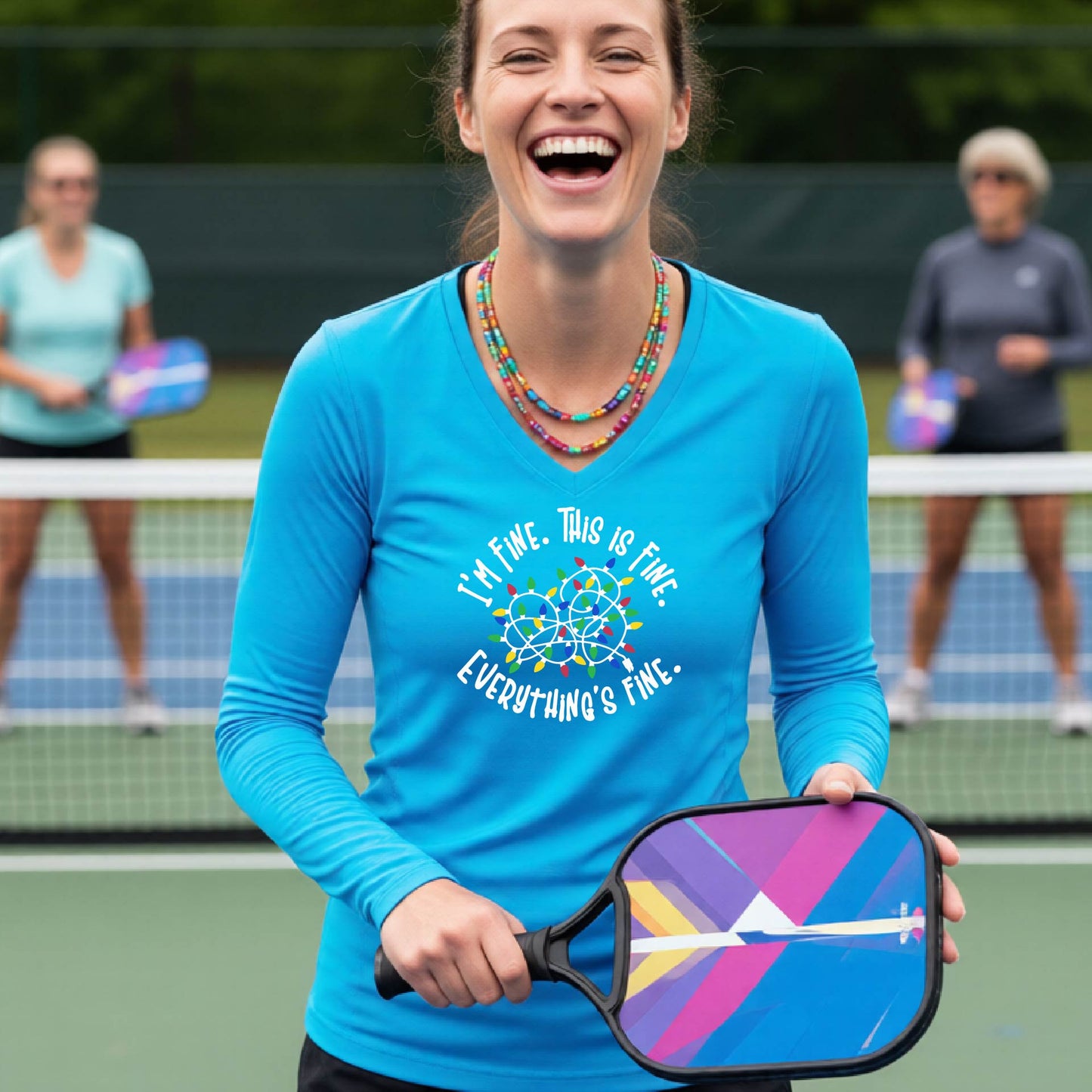 A woman laughing on a pickleball court wearing a bright light blue long-sleeve shirt with a white text graphic and a colorful dreamcatcher design that reads 'I'M FINE. THIS IS FINE.  EVERYTHING'S FINE.'