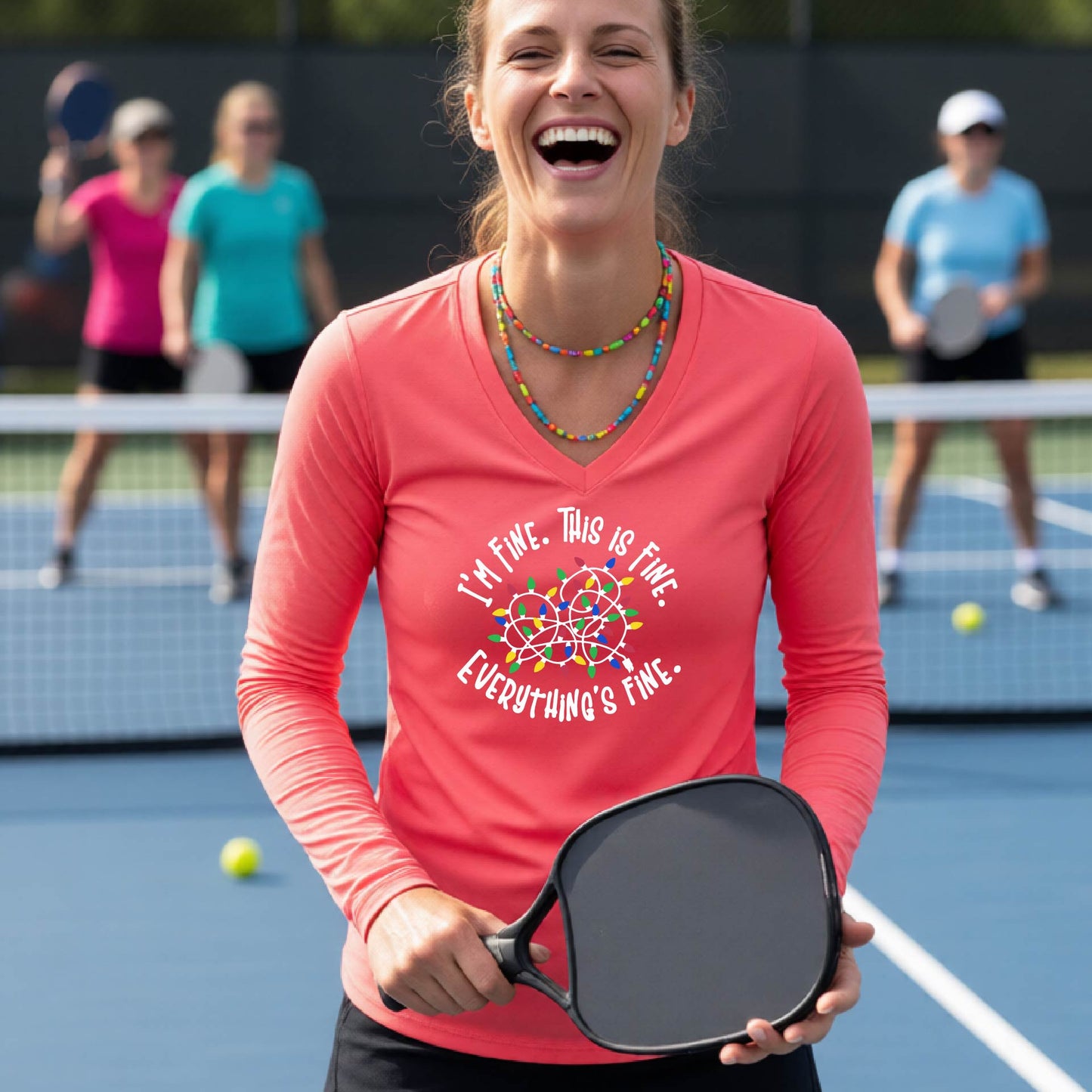 A woman laughing on a pickleball court wearing a bright coral pink long-sleeve shirt with a white text graphic and a colorful dreamcatcher design that reads 'I'M FINE. THIS IS FINE.  EVERYTHING'S FINE.'