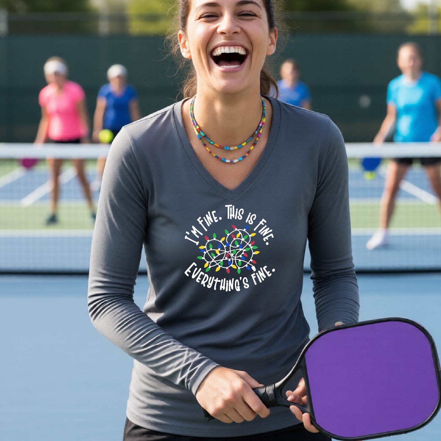 A woman laughing on a pickleball court wearing a dark gray long-sleeve shirt with a white text graphic and a colorful dreamcatcher design that reads 'I'M FINE. THIS IS FINE.  EVERYTHING'S FINE.'