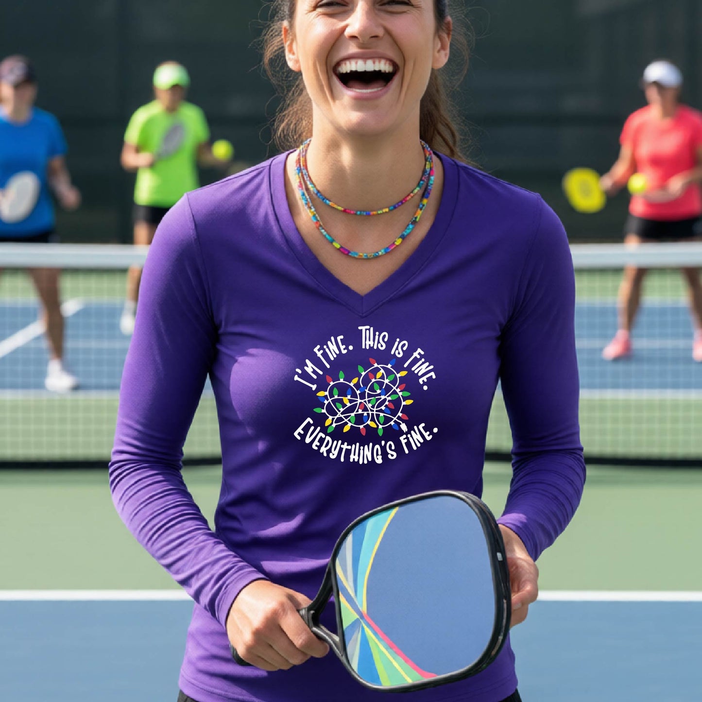 A woman laughing on a pickleball court wearing a purple long-sleeve shirt with a white text graphic and a colorful dreamcatcher design that reads 'I'M FINE. THIS IS FINE.  EVERYTHING'S FINE.'