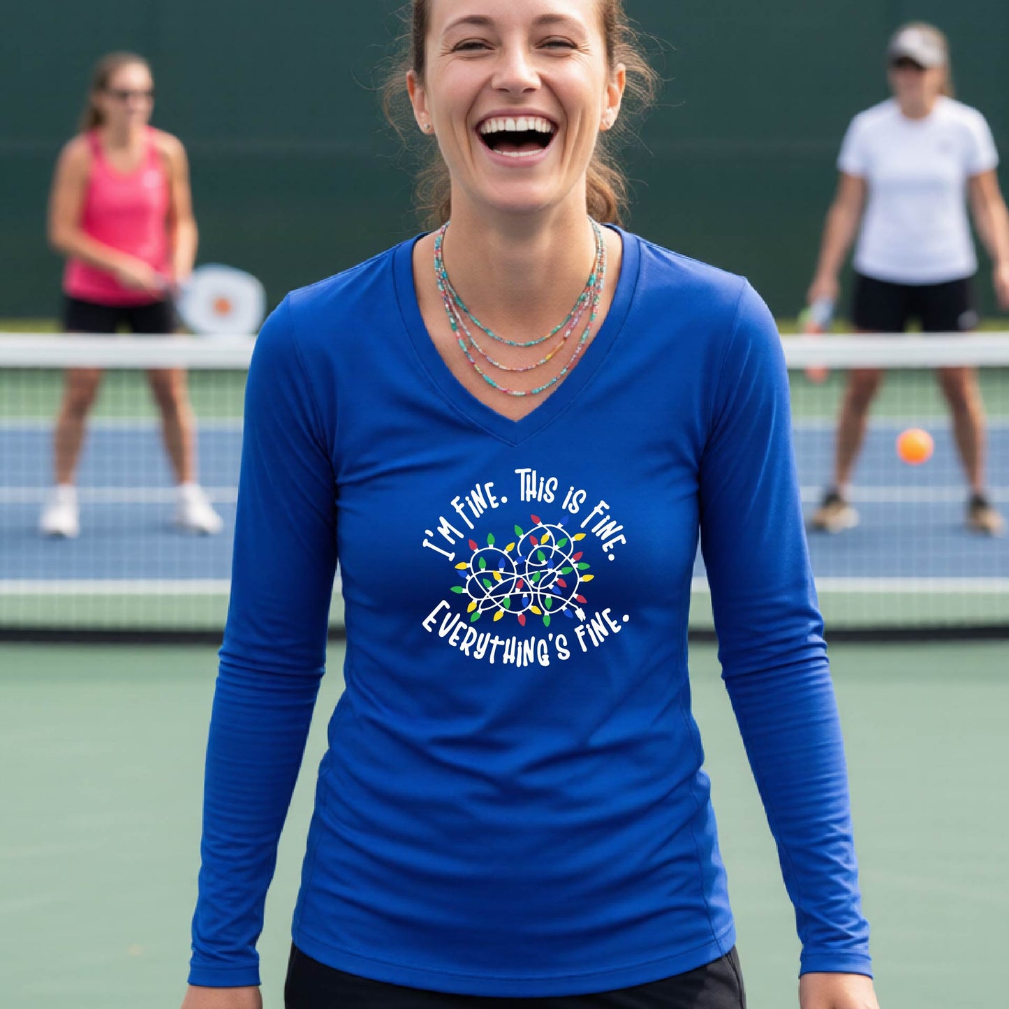 A woman laughing on a pickleball court wearing a royal blue long-sleeve shirt with a white text graphic and a colorful dreamcatcher design that reads 'I'M FINE. THIS IS FINE.  EVERYTHING'S FINE.'