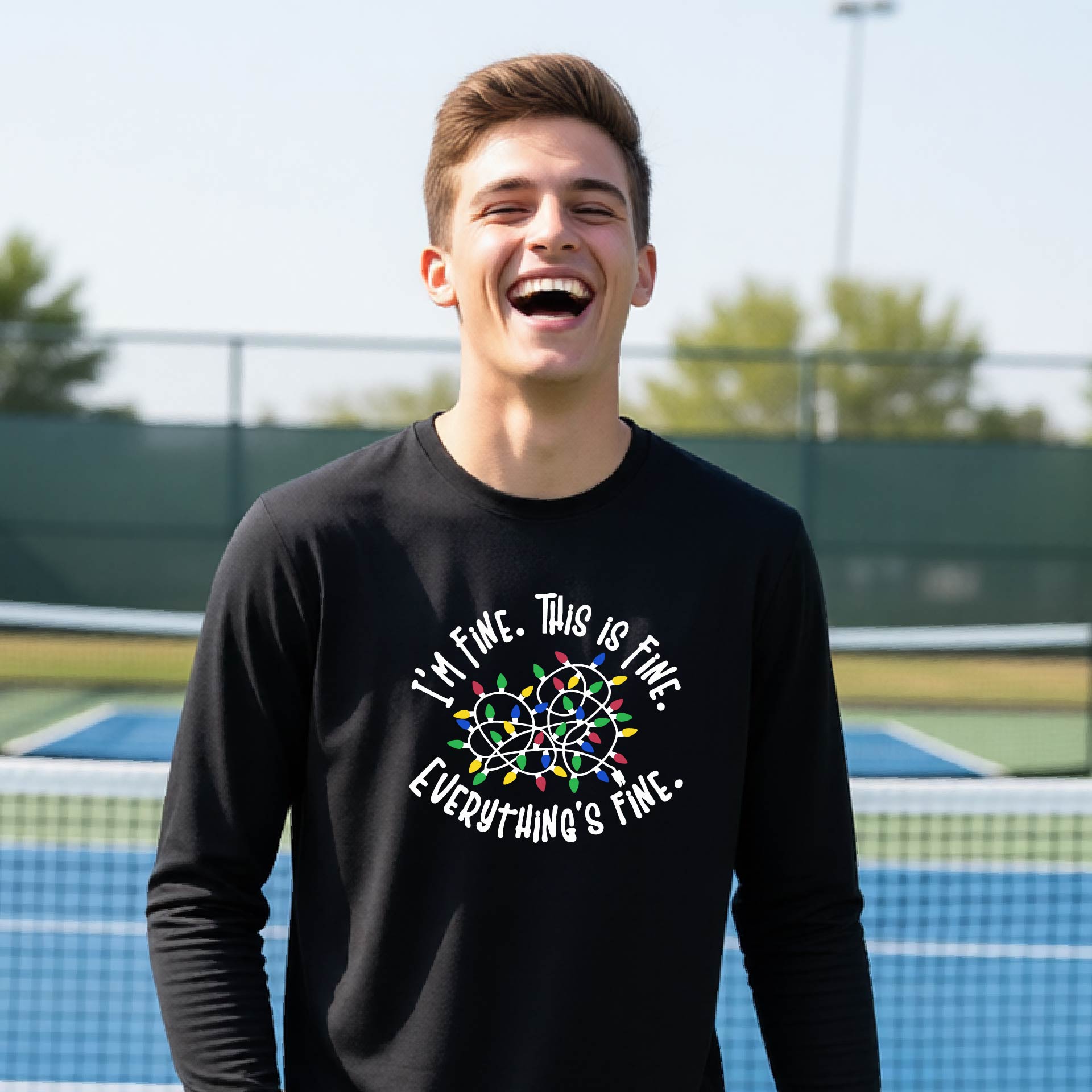 A young adult laughing on a pickleball court wearing a black long-sleeve t-shirt with a white text graphic and a colorful string of lights design that reads 'I'M FINE. THIS IS FINE. EVERYTHING'S FINE.