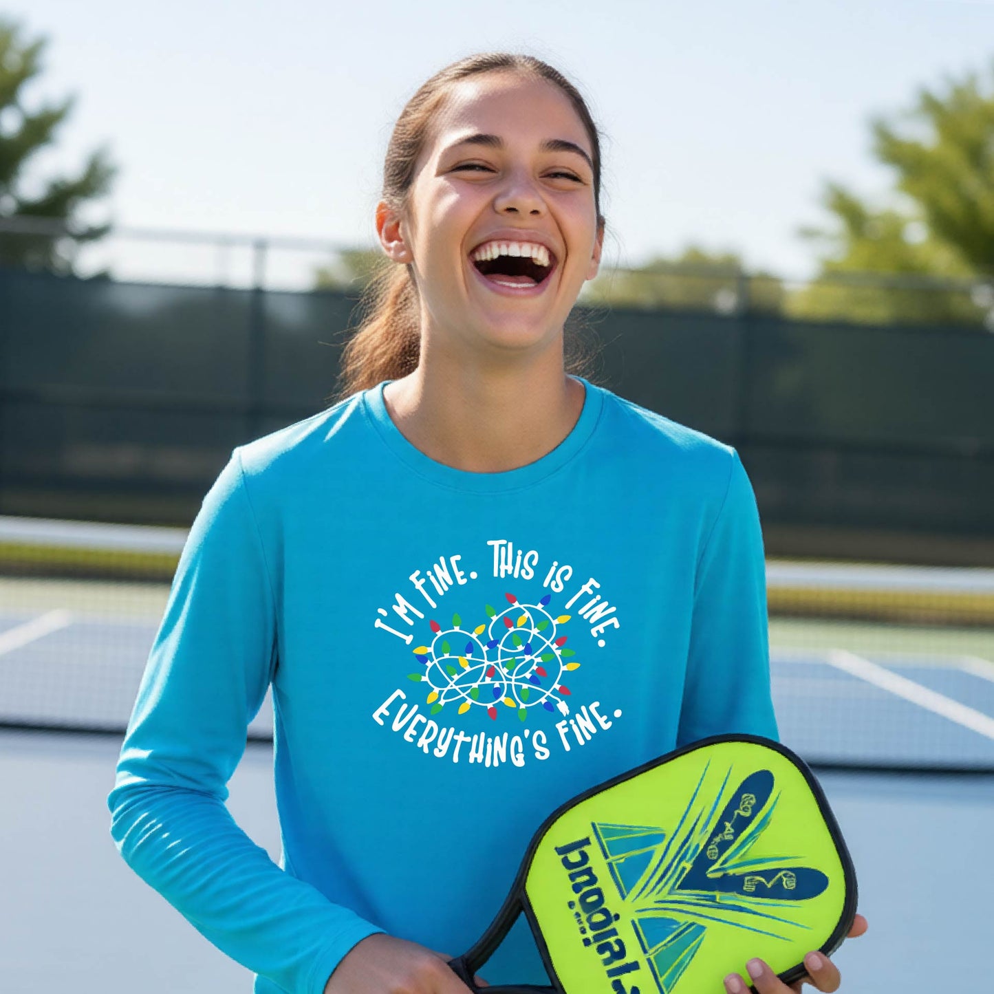A young adult laughing on a pickleball court wearing a bright blue long-sleeve t-shirt with a white text graphic and a colorful string of lights design that reads 'I'M FINE. THIS IS FINE. EVERYTHING'S FINE.