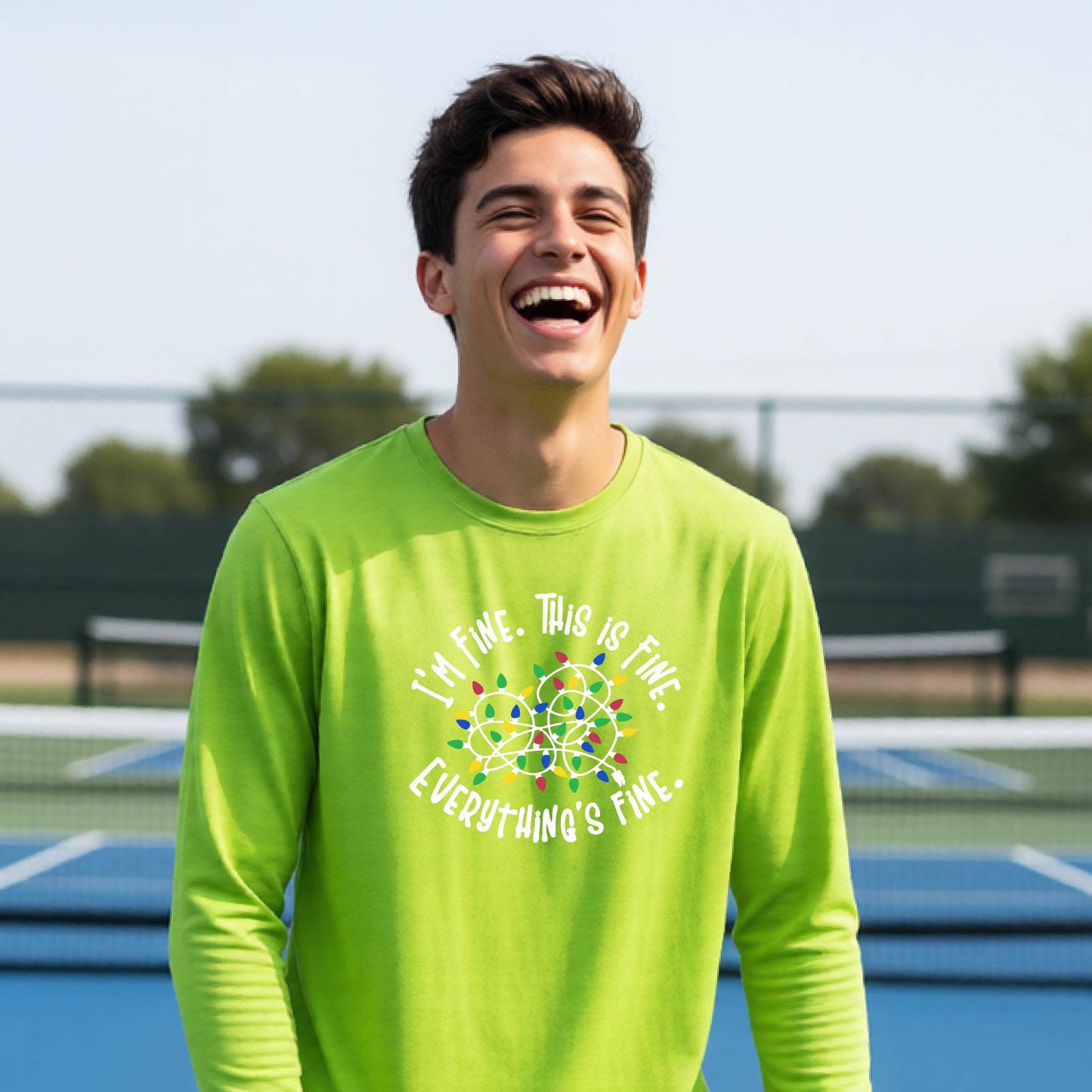 A young adult laughing on a pickleball court wearing a bright lime green  long-sleeve t-shirt with a white text graphic and a colorful string of lights design that reads 'I'M FINE. THIS IS FINE. EVERYTHING'S FINE.
