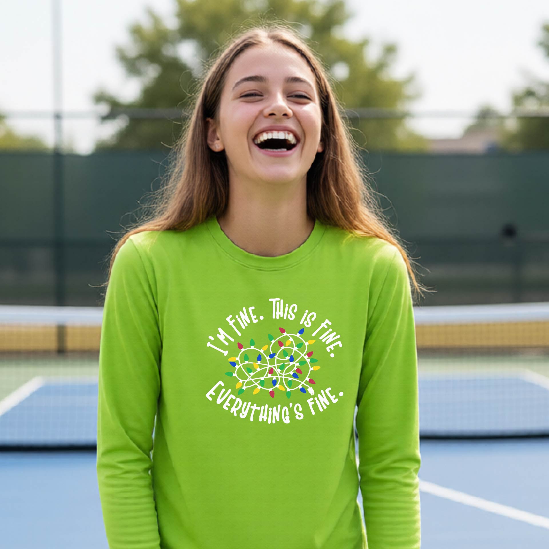 A young adult laughing on a pickleball court wearing a bright lime green long-sleeve t-shirt with a white text graphic and a colorful string of lights design that reads 'I'M FINE. THIS IS FINE. EVERYTHING'S FINE.