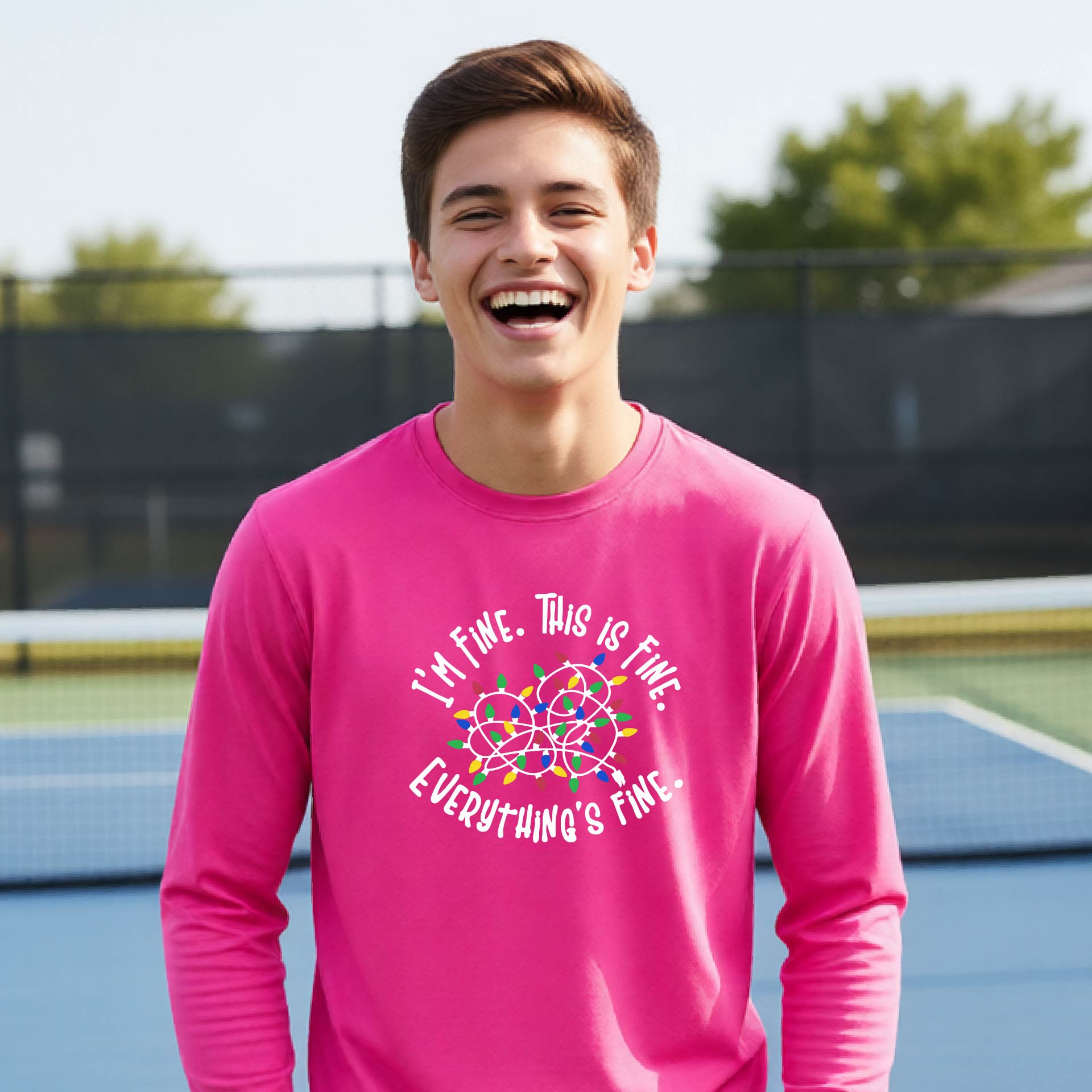 A young adult laughing on a pickleball court wearing a bright pink long-sleeve t-shirt with a white text graphic and a colorful string of lights design that reads 'I'M FINE. THIS IS FINE. EVERYTHING'S FINE.