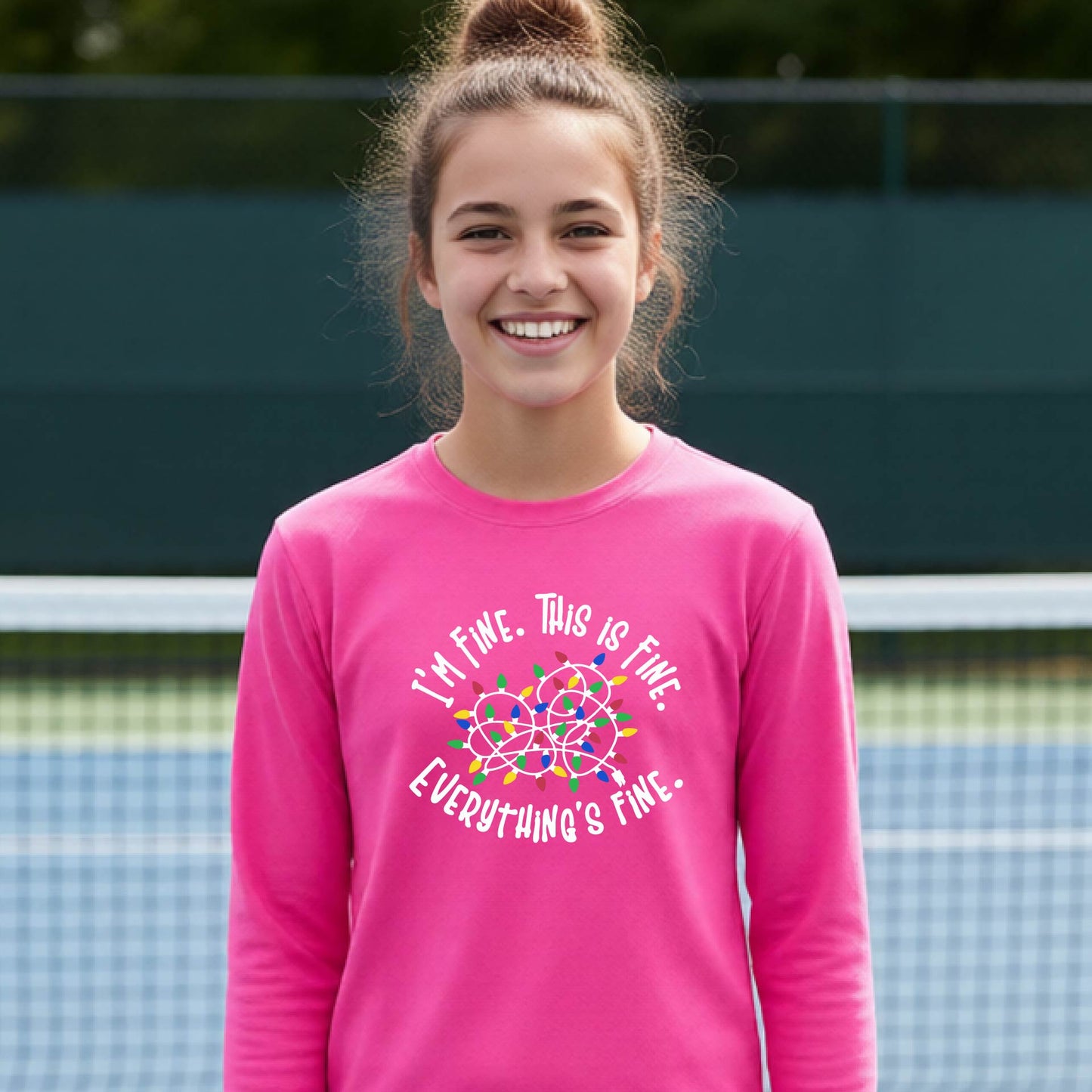 A young adult laughing on a pickleball court wearing a bright pink long-sleeve t-shirt with a white text graphic and a colorful string of lights design that reads 'I'M FINE. THIS IS FINE. EVERYTHING'S FINE.