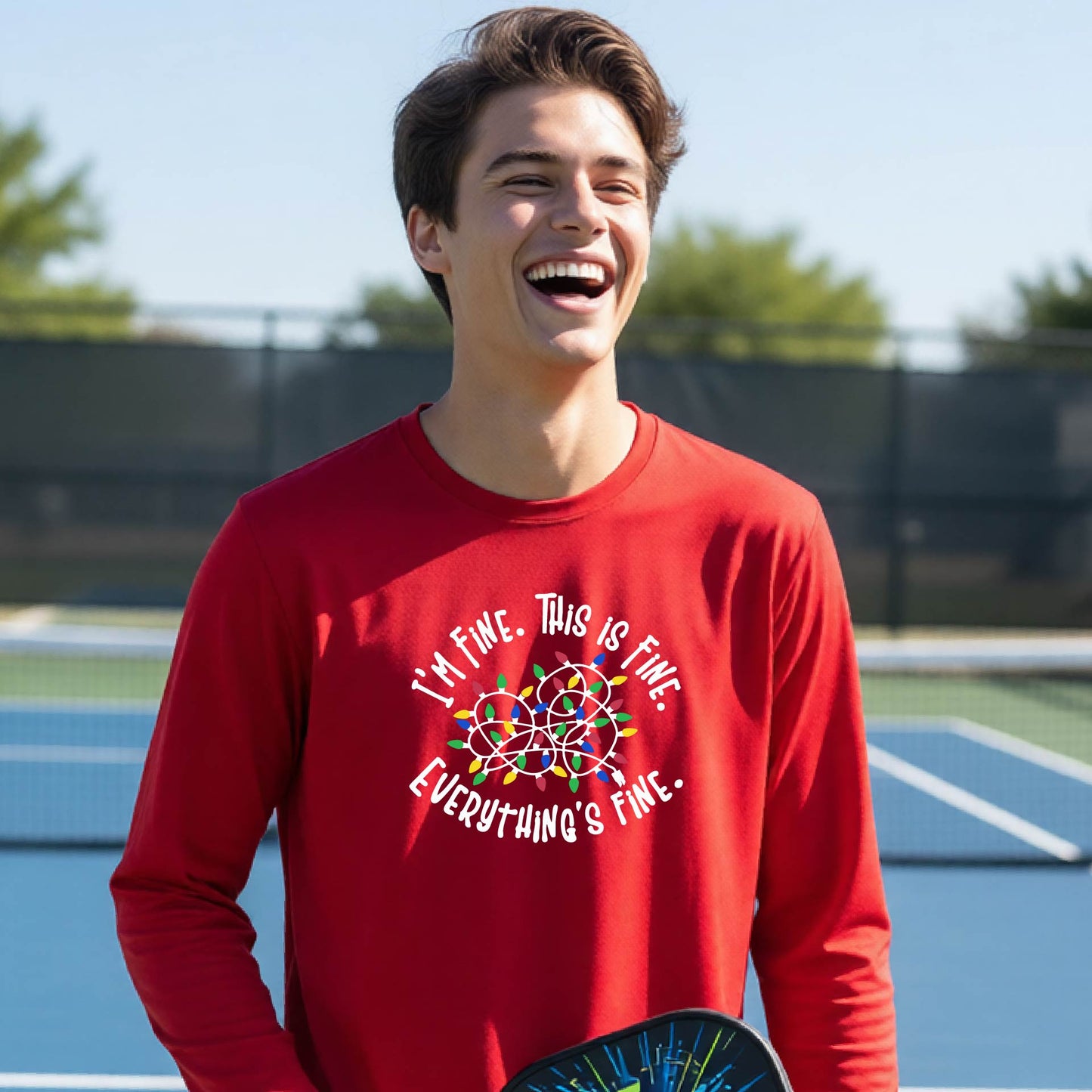 A young adult laughing on a pickleball court wearing a bright red long-sleeve t-shirt with a white text graphic and a colorful string of lights design that reads 'I'M FINE. THIS IS FINE. EVERYTHING'S FINE.