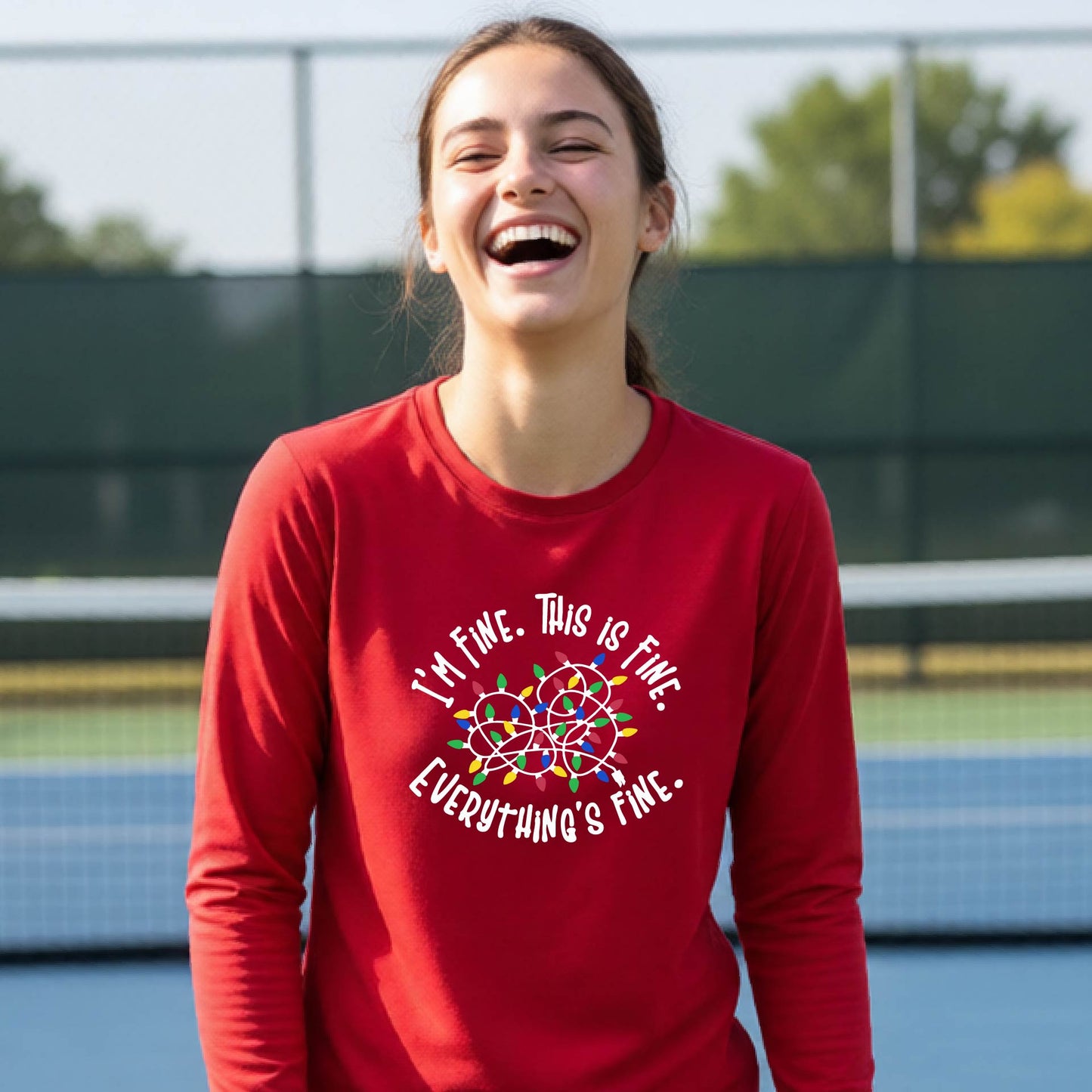 A young adult laughing on a pickleball court wearing a red long-sleeve t-shirt with a white text graphic and a colorful string of lights design that reads 'I'M FINE. THIS IS FINE. EVERYTHING IS FINE.