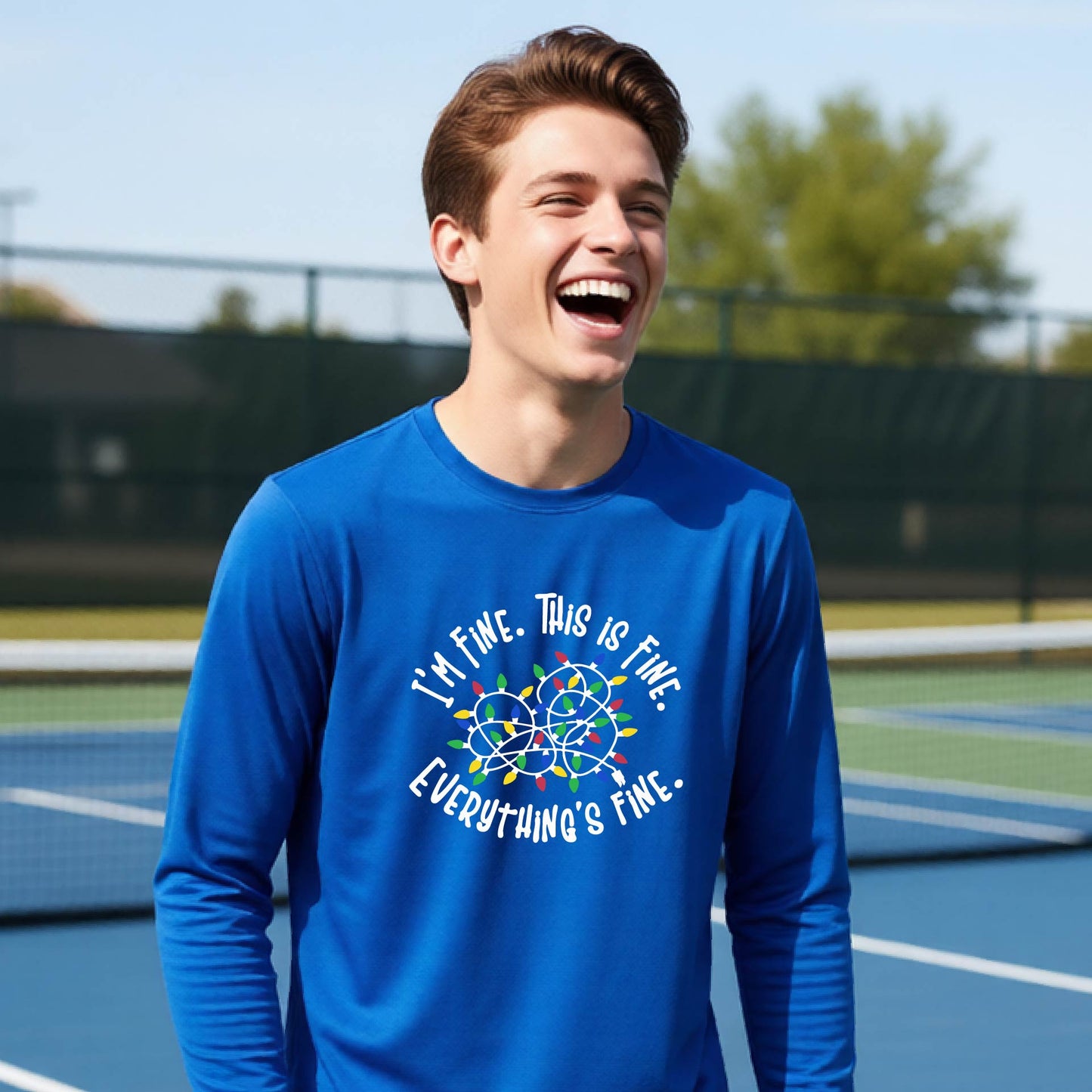 A young adult laughing on a pickleball court wearing a royal blue long-sleeve t-shirt with a white text graphic and a colorful string of lights design that reads 'I'M FINE. THIS IS FINE. EVERYTHING IS FINE.