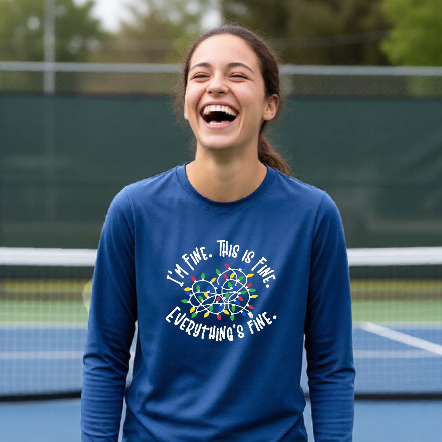 A young adult laughing on a pickleball court wearing a royal blue long-sleeve t-shirt with a white text graphic and a colorful string of lights design that reads 'I'M FINE. THIS IS FINE. EVERYTHING'S FINE.