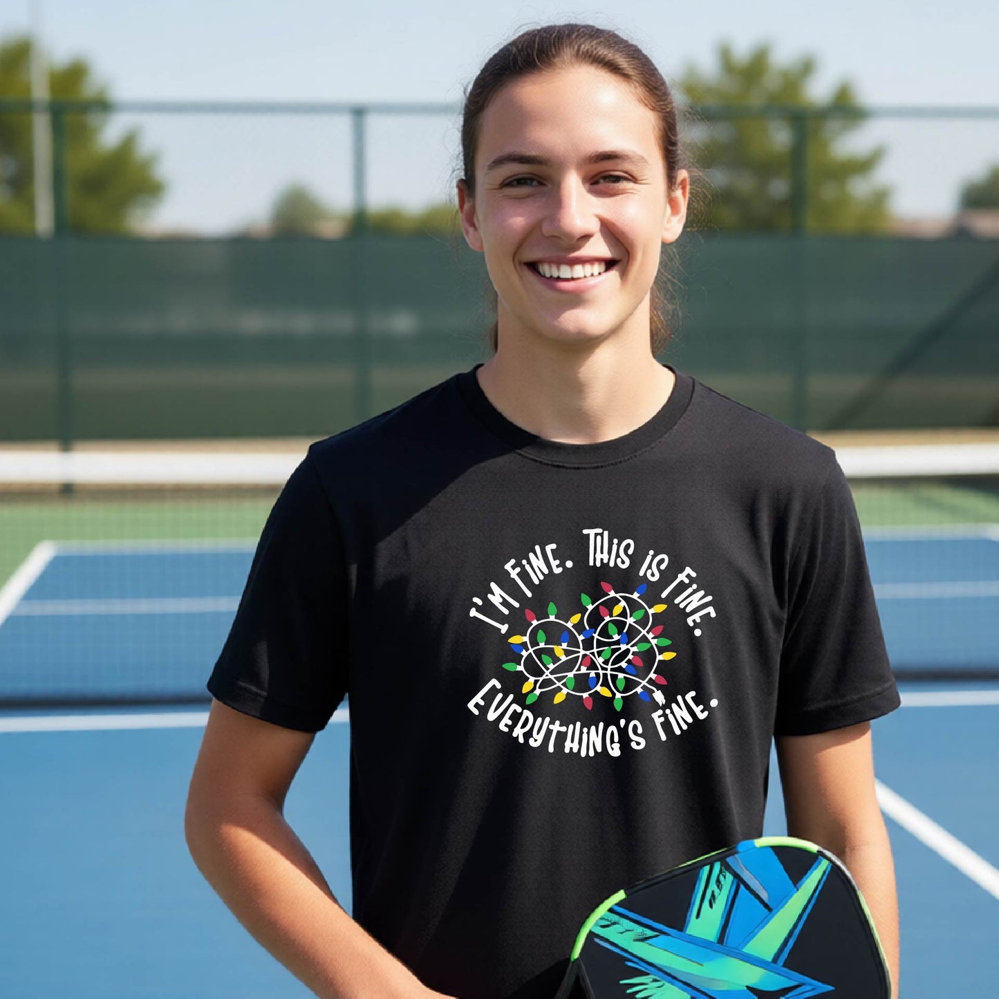 A young adult laughing on a pickleball court wearing a black short-sleeve t-shirt with a white text graphic and a colorful string of lights design that reads 'I'M FINE. THIS IS FINE. EVERYTHING'S FINE.