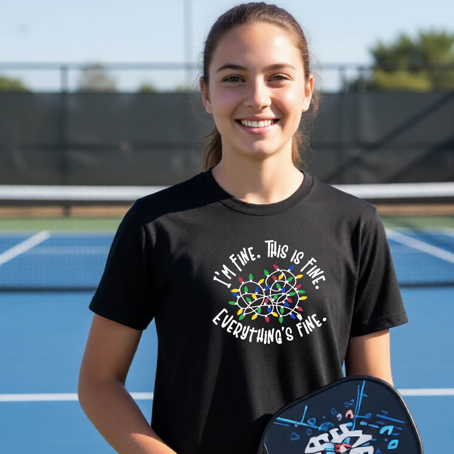 A young adult laughing on a pickleball court wearing a black short-sleeve t-shirt with a white text graphic and a colorful string of lights design that reads 'I'M FINE. THIS IS FINE. EVERYTHING'S FINE.THIS IS FIVE.