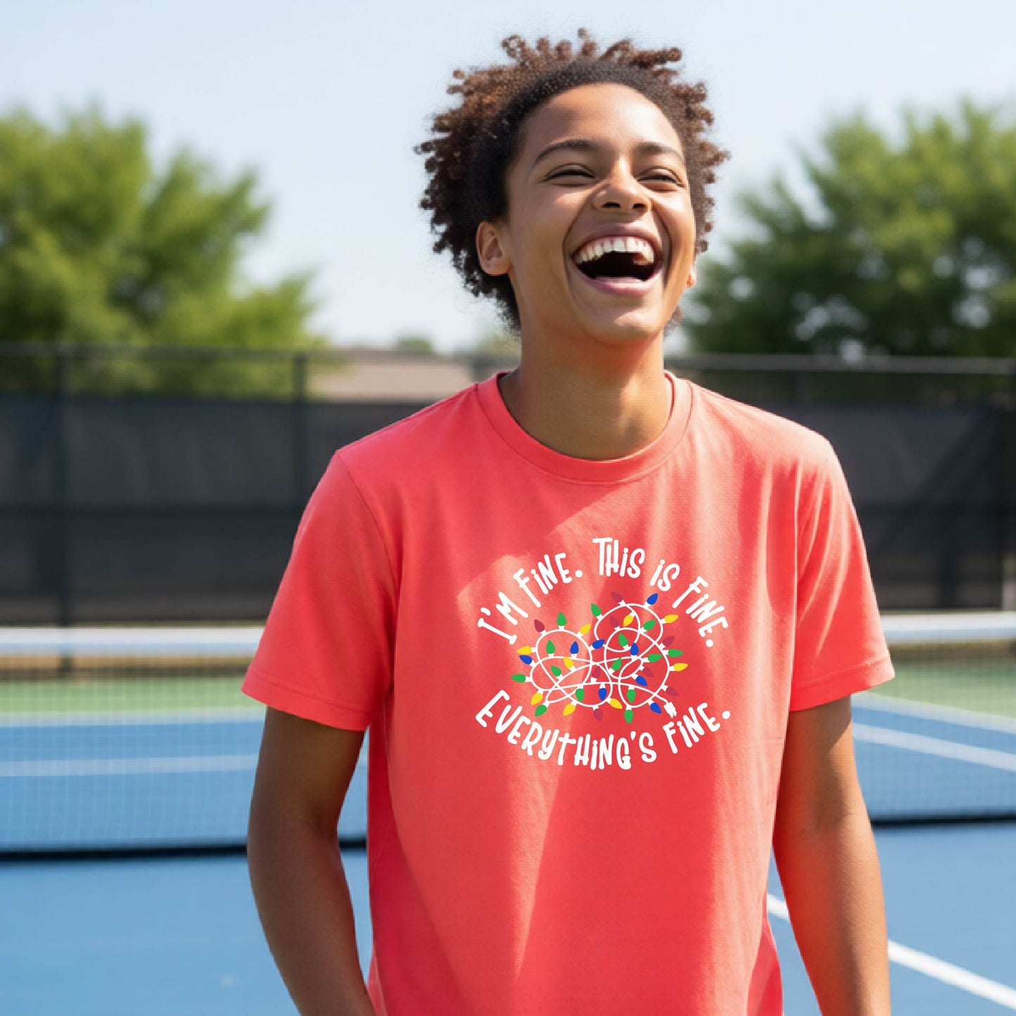 A young adult laughing on a pickleball court wearing a coral-colored short-sleeve t-shirt with a white text graphic and a colorful string of lights design that reads 'I'M FINE. THIS IS FINE. EVERYTHING'S FINE.