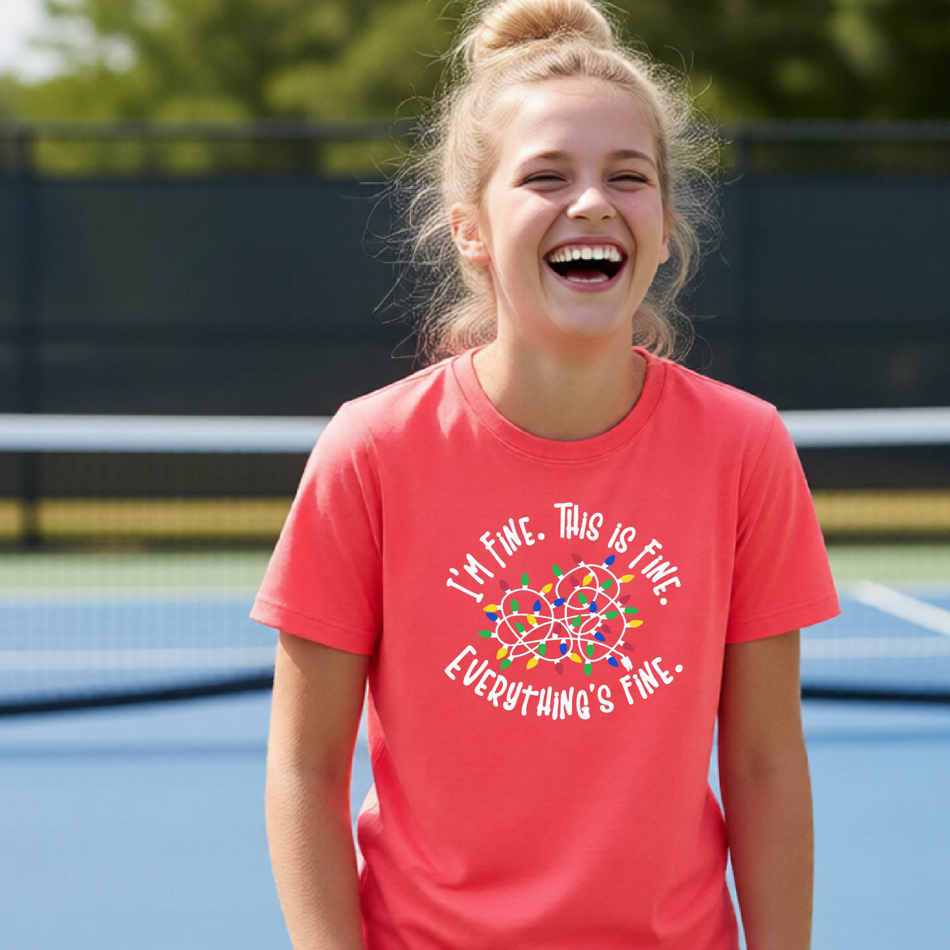 A young adult laughing on a pickleball court wearing a bright blue short-sleeve t-shirt with a white text graphic and a colorful string of lights design that reads 'I'M FINE. THIS IS FINE. EVERYTHING'S FINE.
