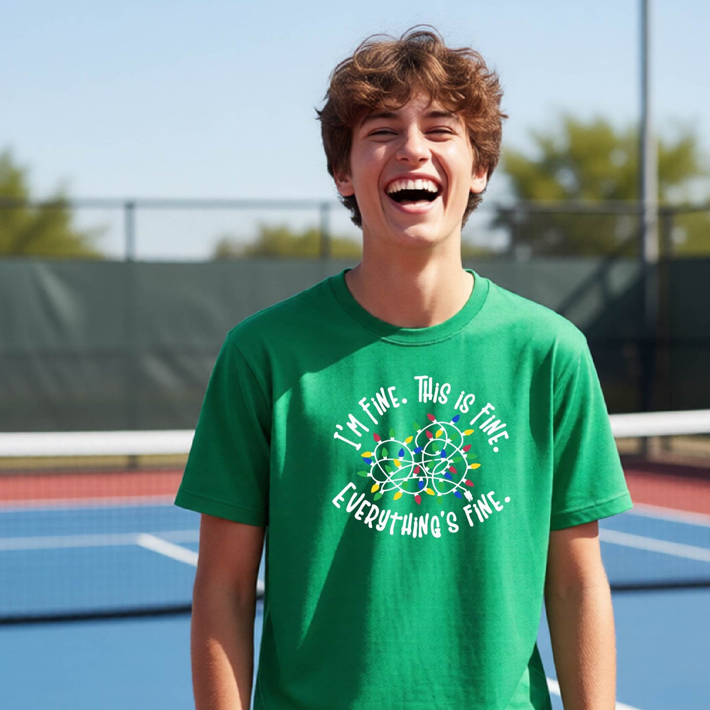A young adult laughing on a pickleball court wearing a green short-sleeve t-shirt with a white text graphic and a colorful string of lights design that reads 'I'M FINE. THIS IS FINE. EVERYTHING'S FINE.