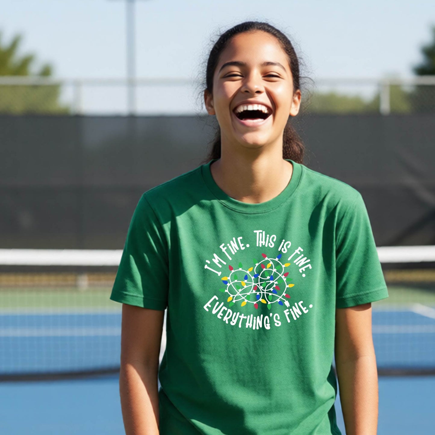 A young adult laughing on a pickleball court wearing a green short-sleeve t-shirt with a white text graphic and a colorful string of lights design that reads 'I'M FINE. THIS IS FINE. EVERYTHING'S FINE.