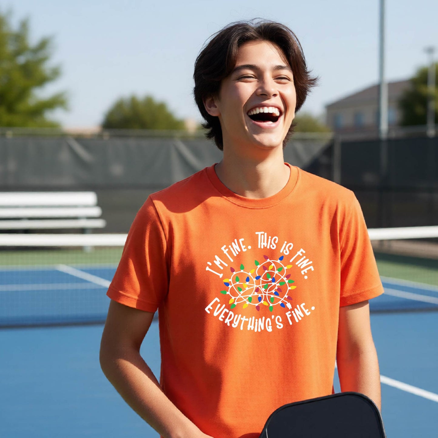 A young adult laughing on a pickleball court wearing an orange short-sleeve t-shirt with a white text graphic and a colorful string of lights design that reads 'I'M FINE. THIS IS FINE. EVERYTHING'S FINE.