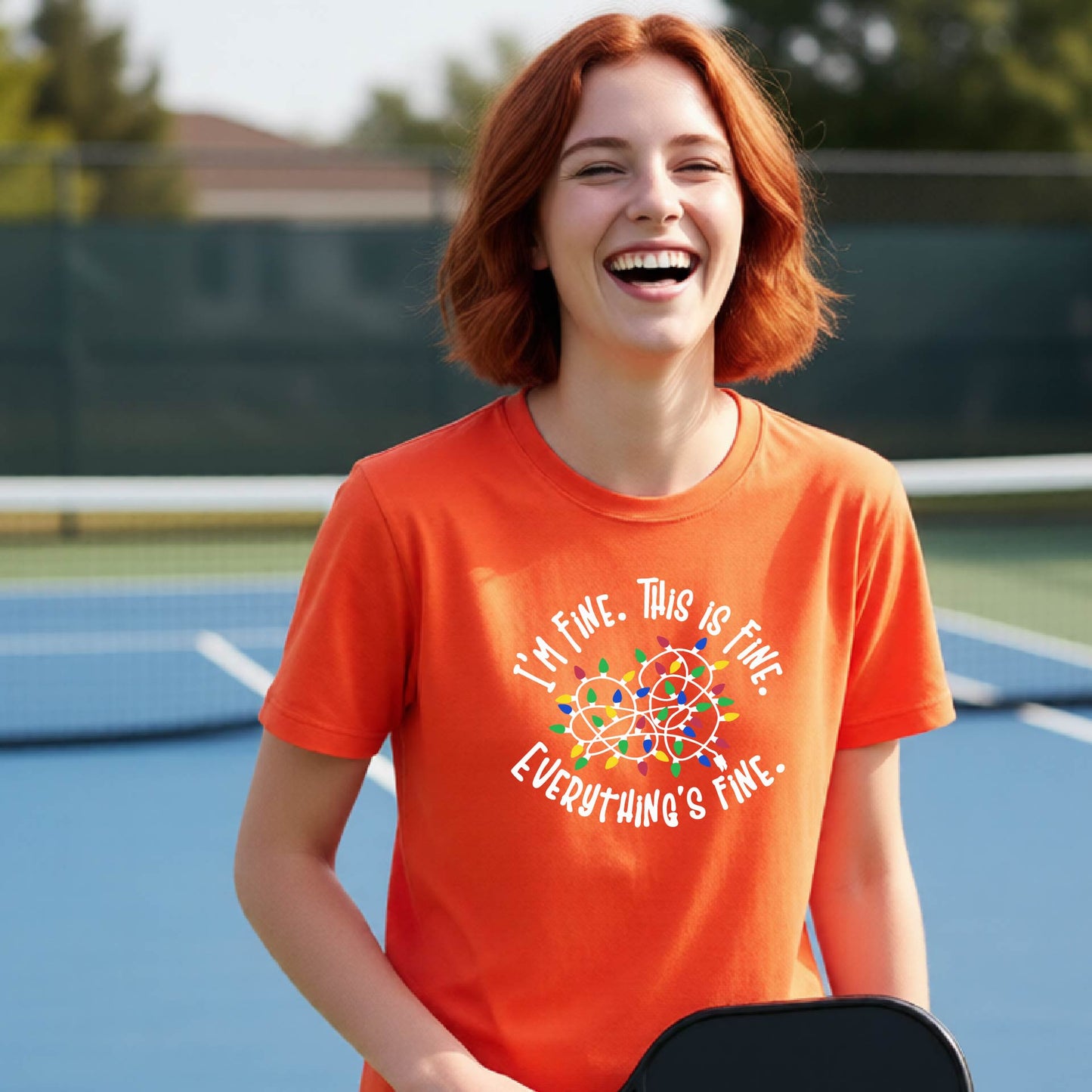 A young adult laughing on a pickleball court wearing an orange short-sleeve t-shirt with a white text graphic and a colorful string of lights design that reads 'I'M FINE. THIS IS FINE. EVERYTHING'S FINE.