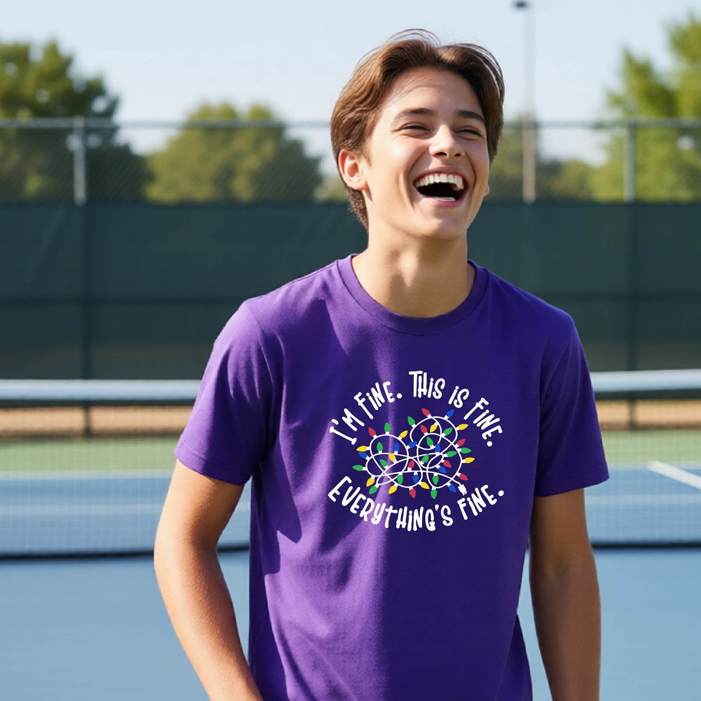 A young adult laughing on a pickleball court wearing a purple short-sleeve t-shirt with a white text graphic and a colorful string of lights design that reads 'I'M FINE. THIS IS FINE. EVERYTHING'S FINE.