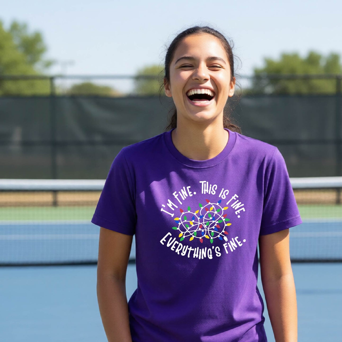 A young adult laughing on a pickleball court wearing a purple short-sleeve t-shirt with a white text graphic and a colorful string of lights design that reads 'I'M FINE. THIS IS FINE. EVERYTHING'S FINE.