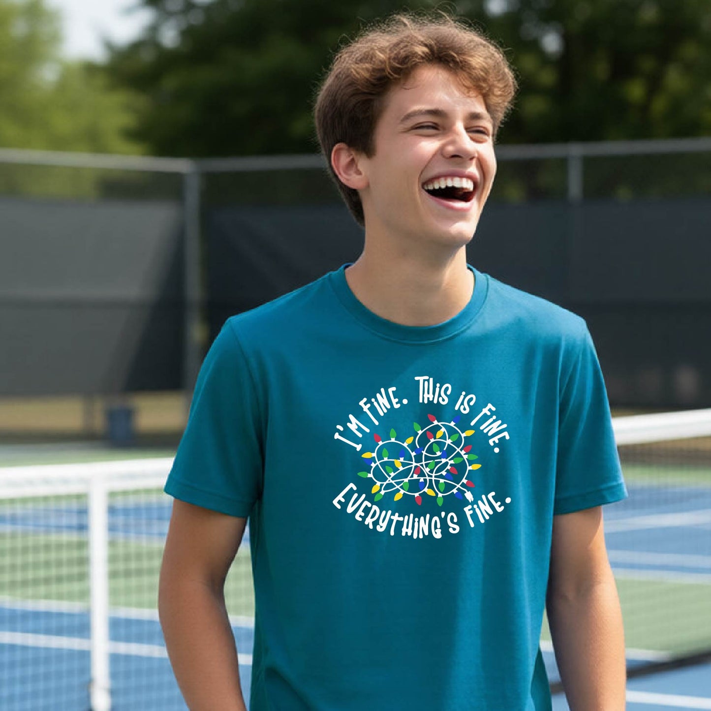A young adult laughing on a pickleball court wearing a teal short-sleeve t-shirt with a white text graphic and a colorful string of lights design that reads 'I'M FINE. THIS IS FINE. EVERYTHING'S FINE.