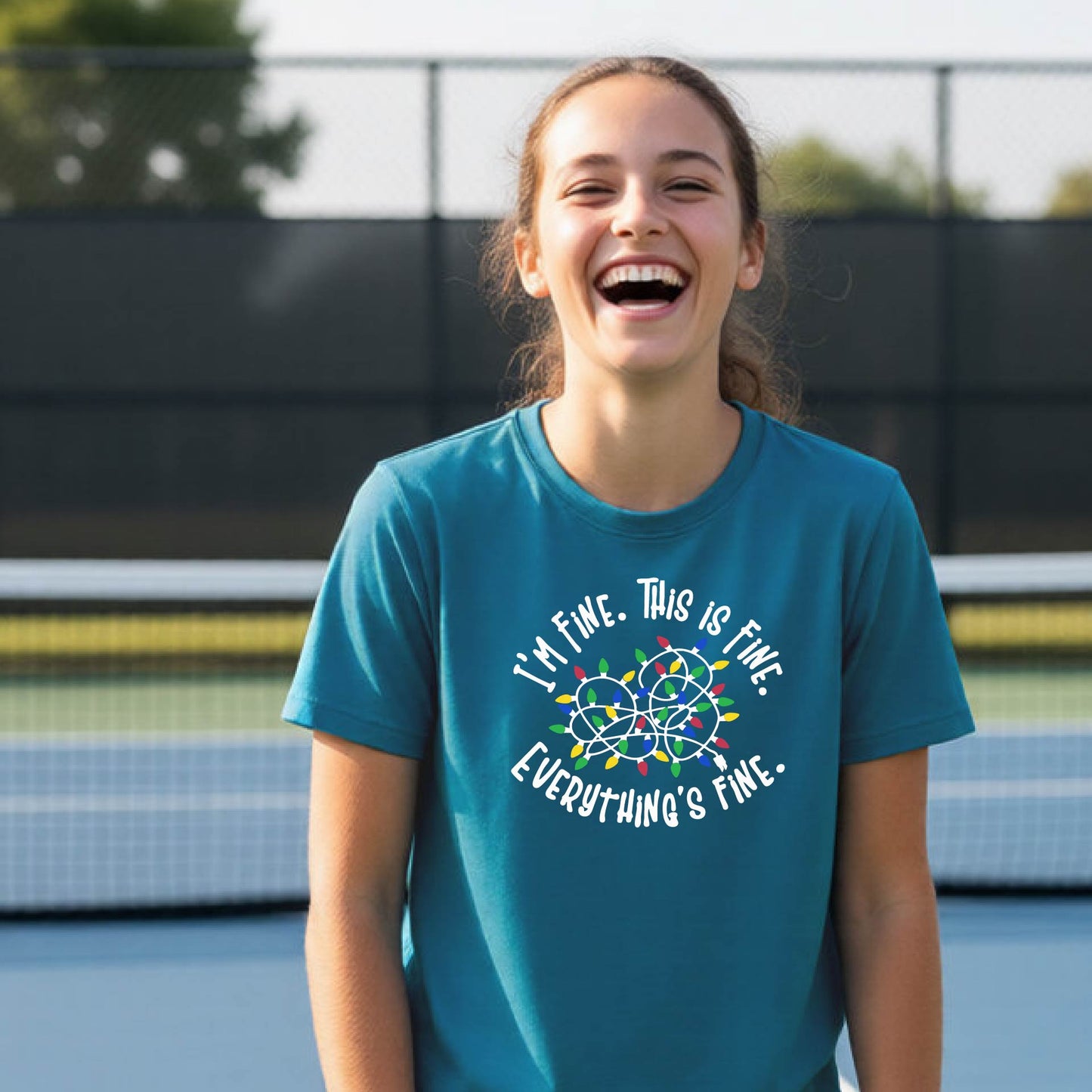 A young adult laughing on a pickleball court wearing a teal short-sleeve t-shirt with a white text graphic and a colorful string of lights design that reads 'I'M FINE. THIS IS FINE. EVERYTHING'S FINE.